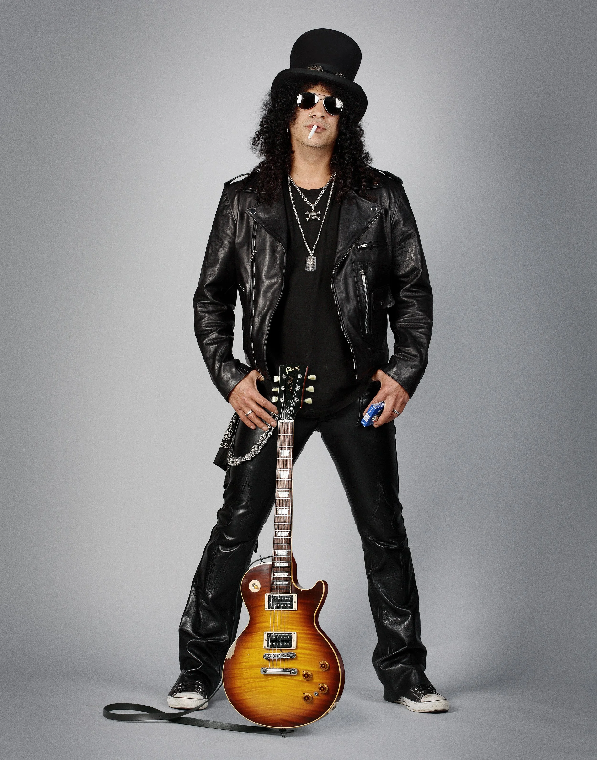 A man dressed in a black leather jacket, black pants, and a black top hat, wearing sunglasses and jewelry, holding a yellow-orange Gibson electric guitar, standing against a gray background.