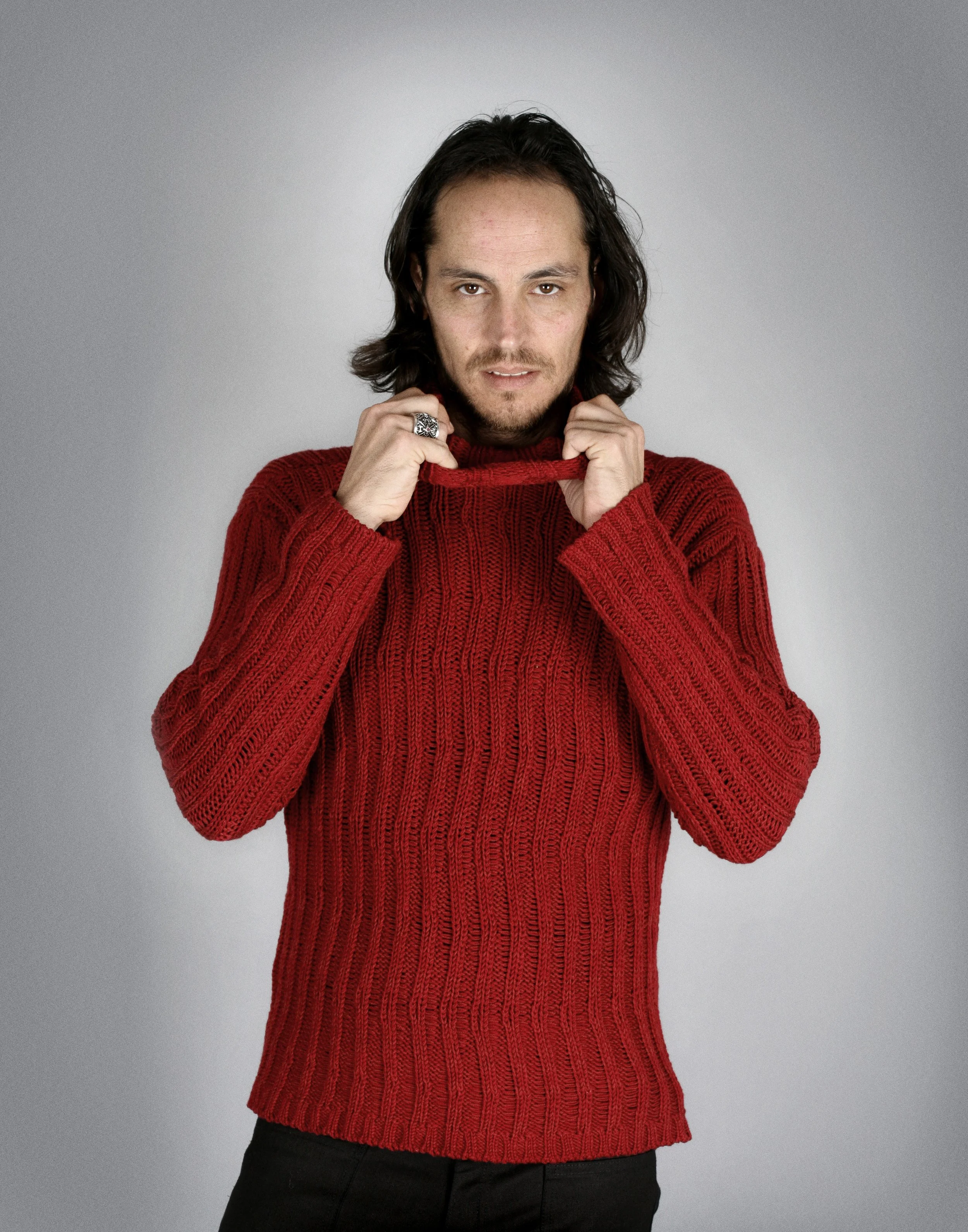 Man with long dark hair wearing a red knitted sweater, holding the collar and looking at the camera against a plain gray background.