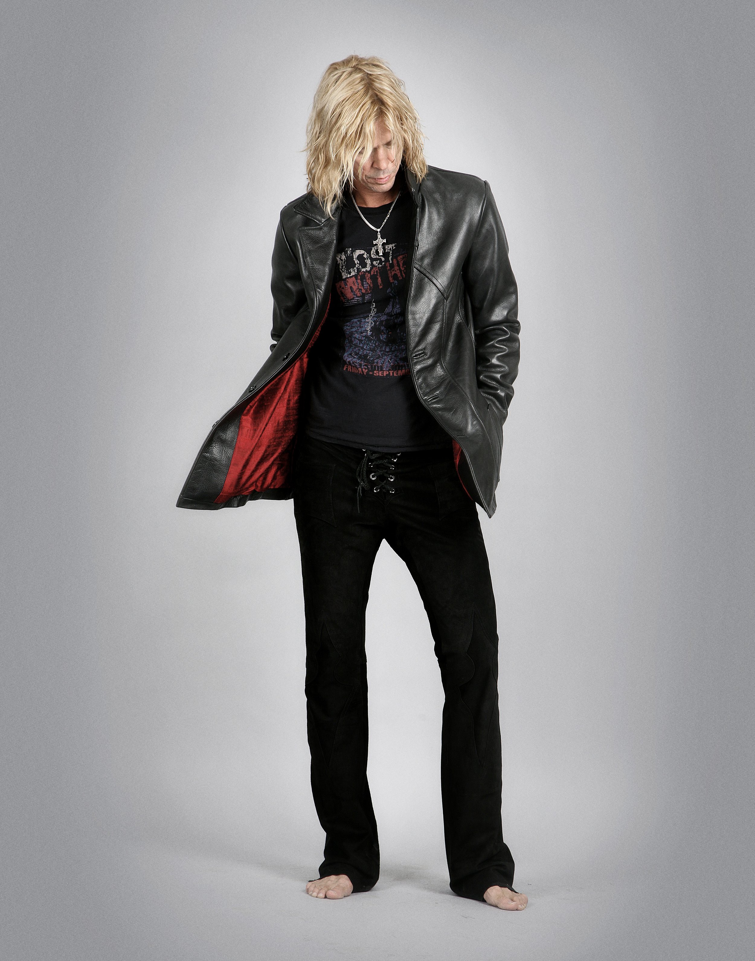 Man with long blond hair wearing a black leather jacket, black graphic t-shirt, black pants, barefoot, against gray background.