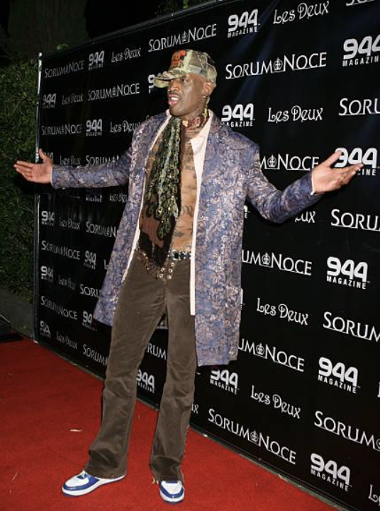 Man posing with arms outstretched at a media event, wearing a patterned blazer, scarf, tan pants, sneakers, and a camo cap, in front of a black step-and-repeat backdrop with logos for 944 Magazine, SorriNoce, and Les Deux.