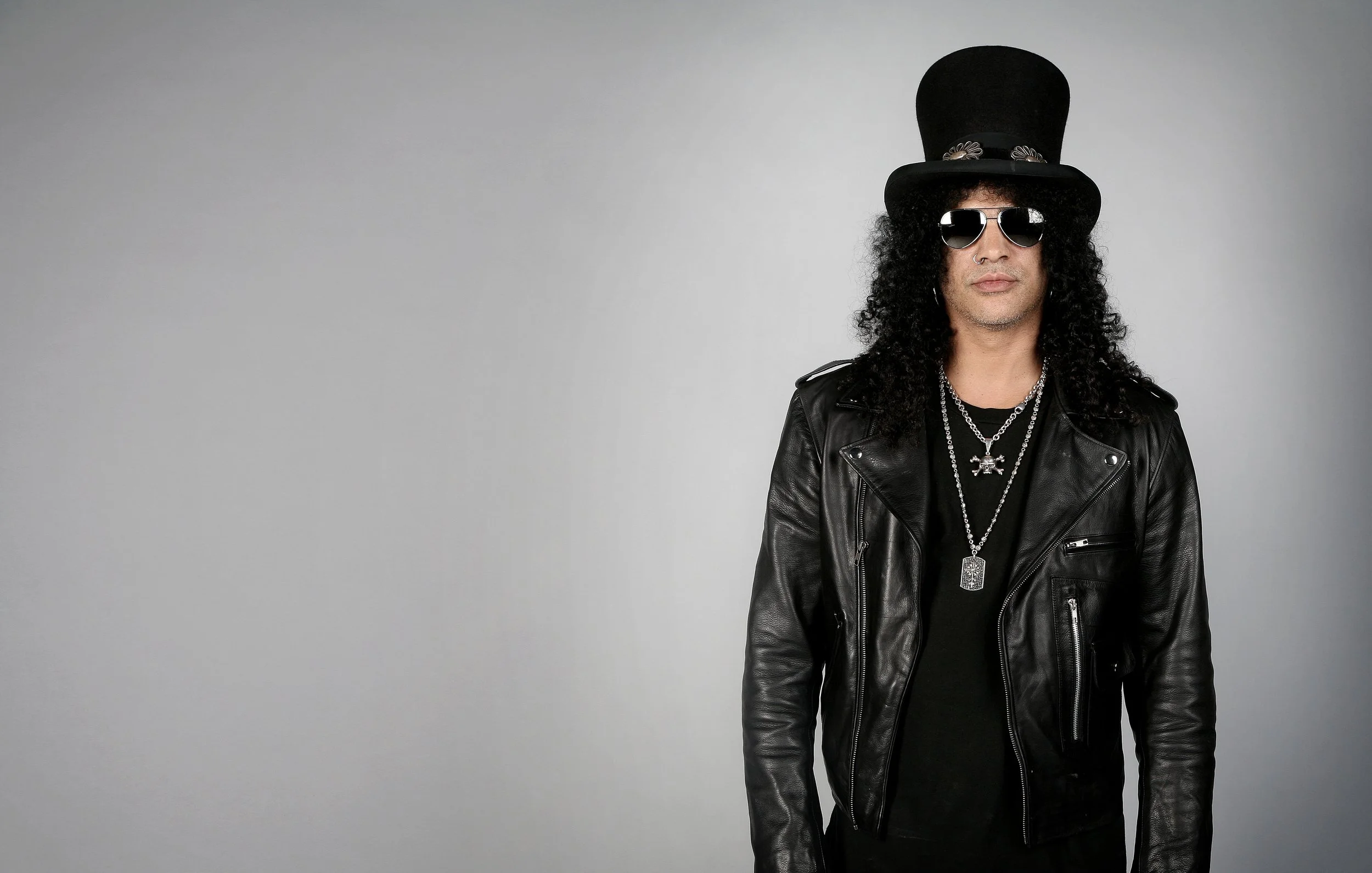 Man wearing black leather jacket, black t-shirt, multiple silver chains, black top hat, and round sunglasses against a plain gray background.