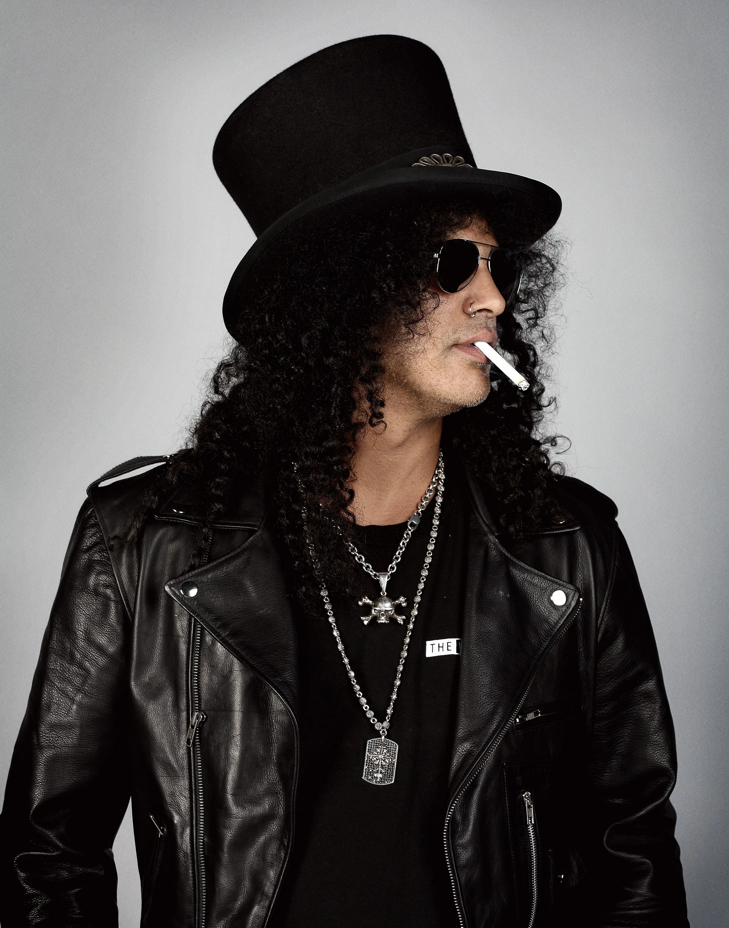 Person with curly hair wearing a black leather jacket, multiple necklaces, black sunglasses, and a tall black hat, holding a cigarette in their mouth, against a plain gray background.