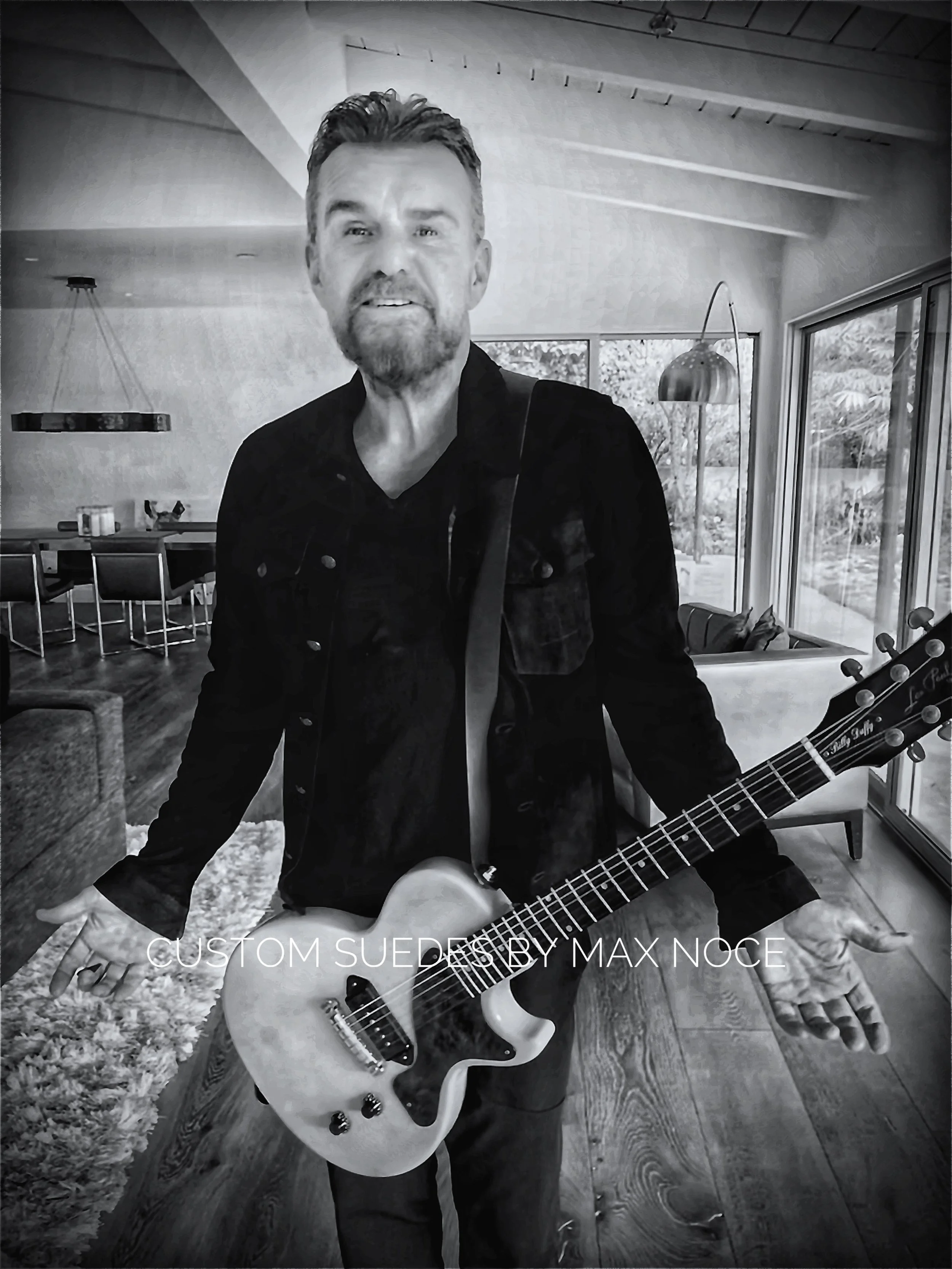 Man with electric guitar standing inside a modern living room with large windows, hardwood floors, and contemporary furniture.
