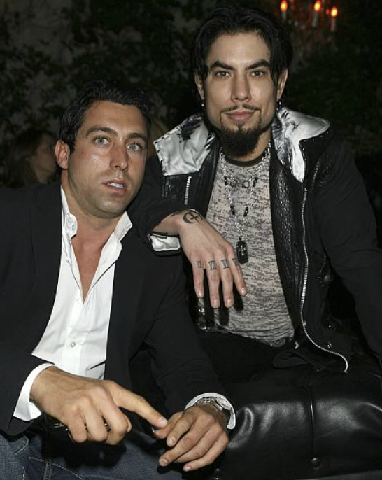 Two men sitting together at a social event, one in a suit and the other in casual attire with tattoos, both looking at the camera.