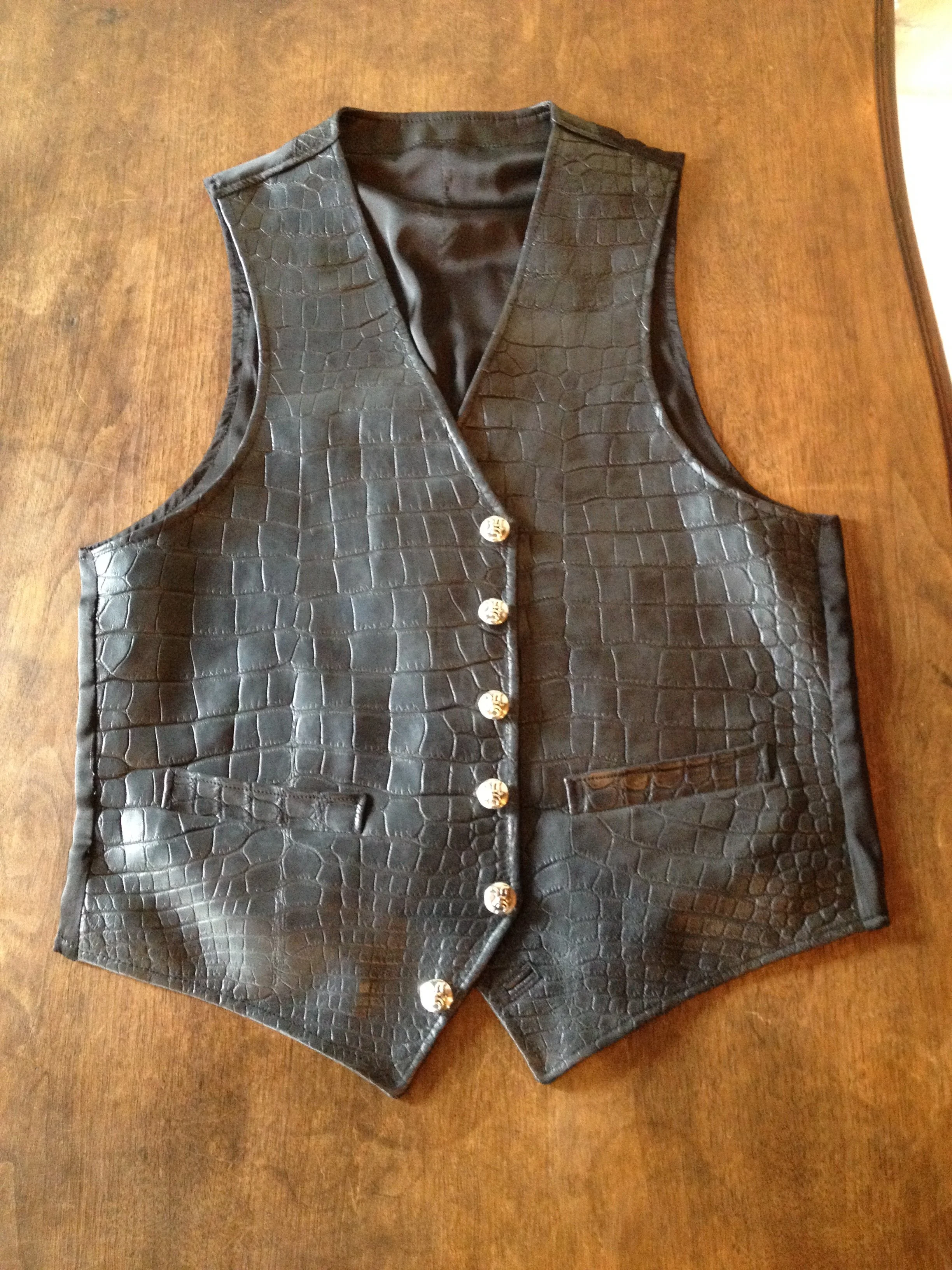 Black sleeveless vest with crocodile pattern, gold buttons, and front pocket, laid on wooden surface.
