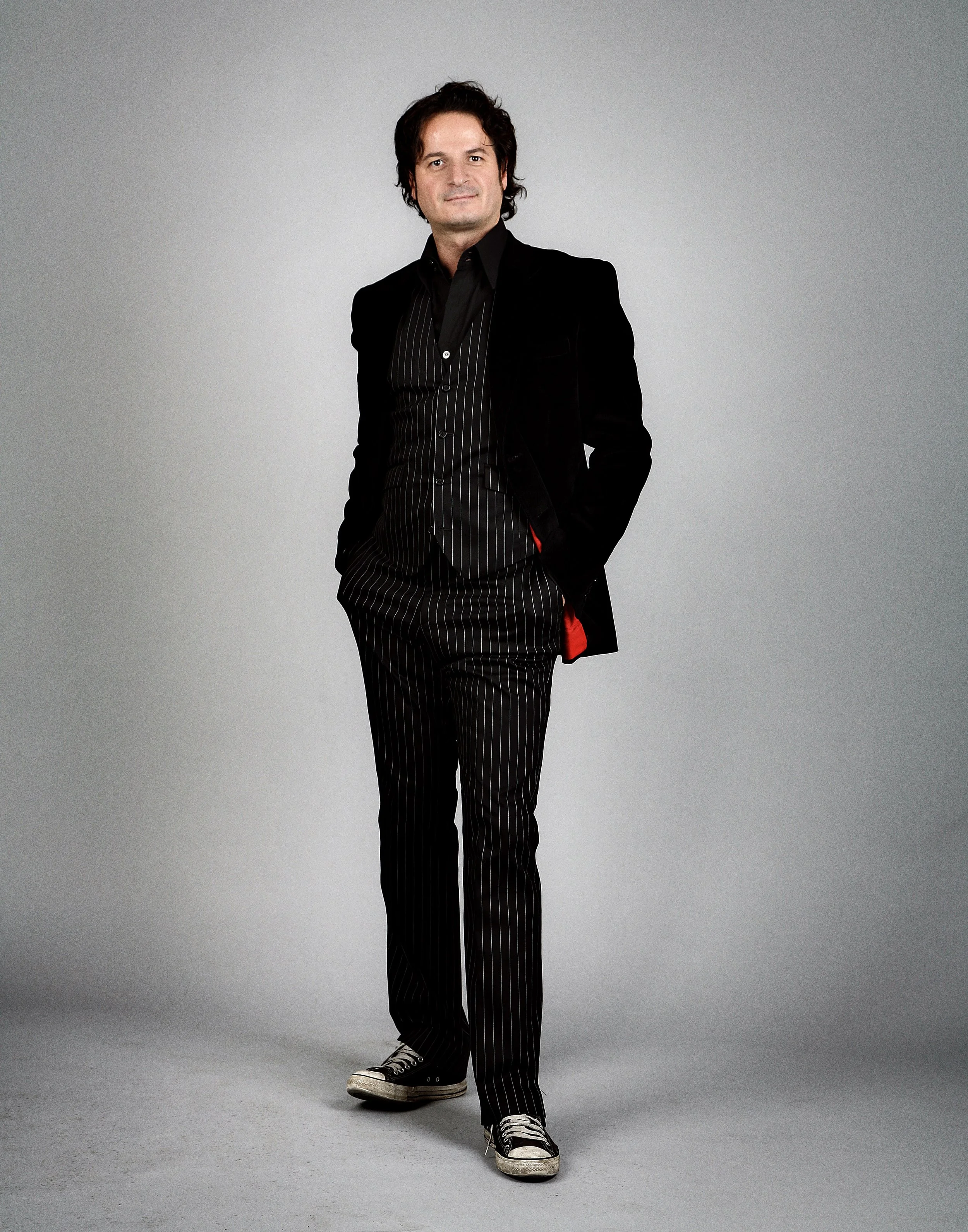 A man with dark, wavy hair wearing a black pinstripe suit, black shirt, and sneakers stands against a plain gray background.