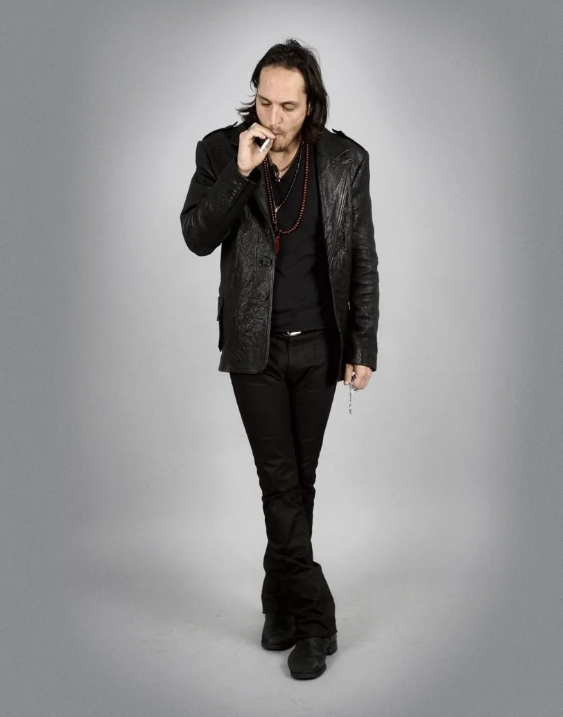 A man with long dark hair and a goatee wearing a black leather jacket, black shirt, black pants, and black shoes, standing against a plain gray background and smoking a cigarette.