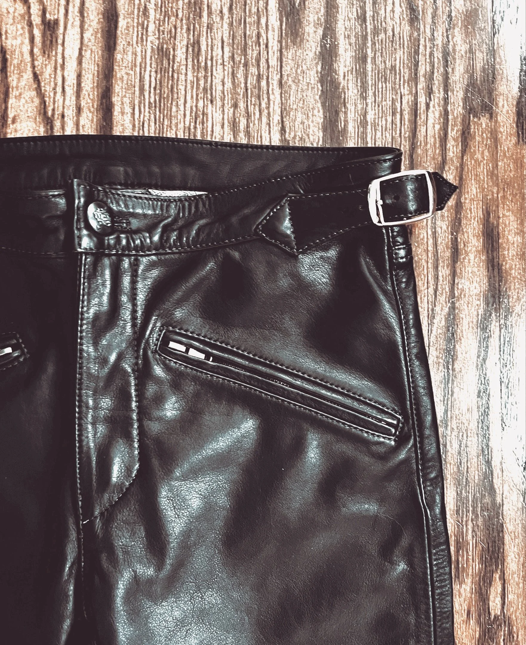 Close-up of black leather pants with zipper pocket, belt loop, and buckle on a wooden surface.