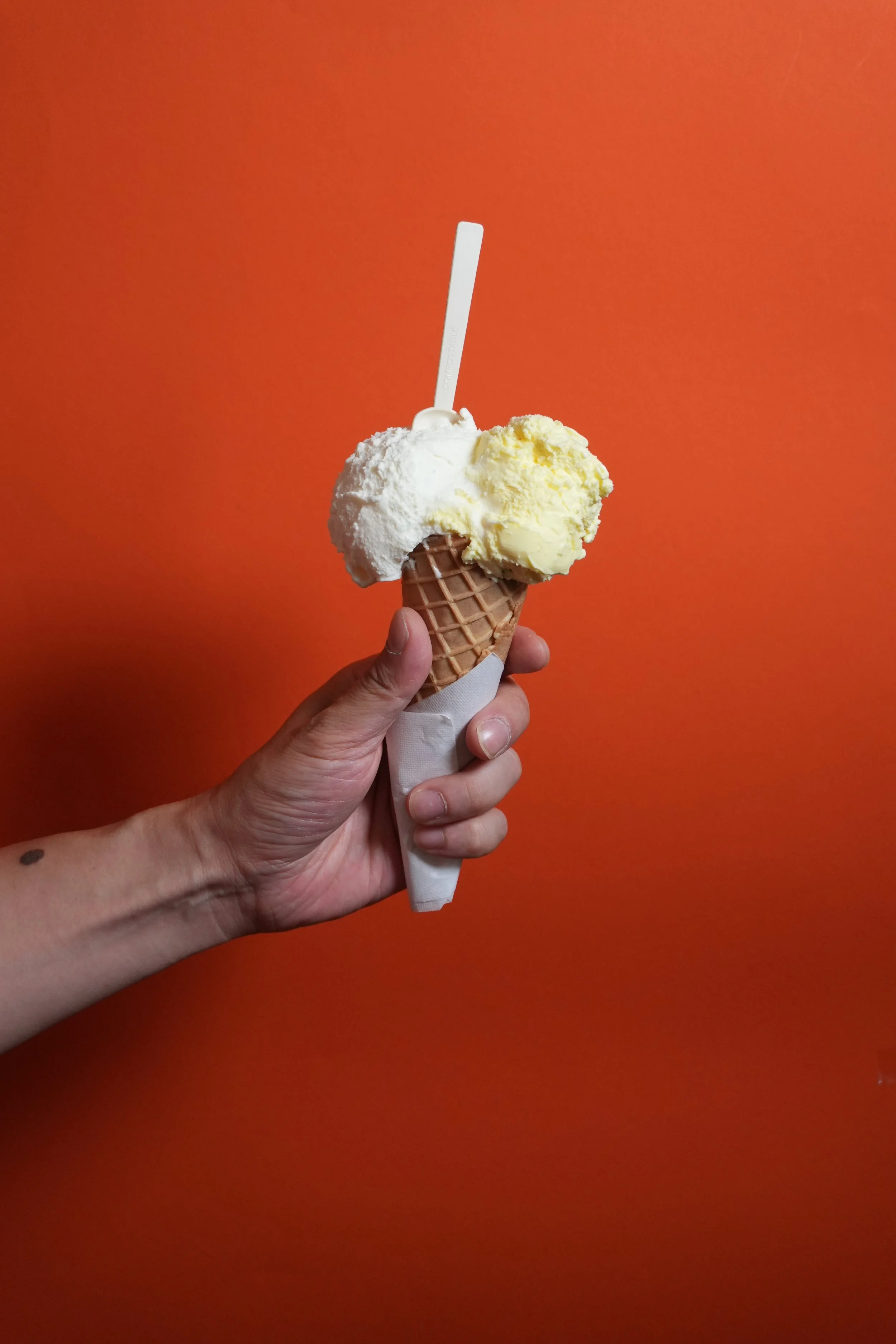 Photo prise par Dreamer's Table avec une main qui tient un cone glacée avec deux boules de glace, une blanche et une jaune, dans un cornet en forme de gaufrette contre un fond orange au magasin La Bise à Lausanne