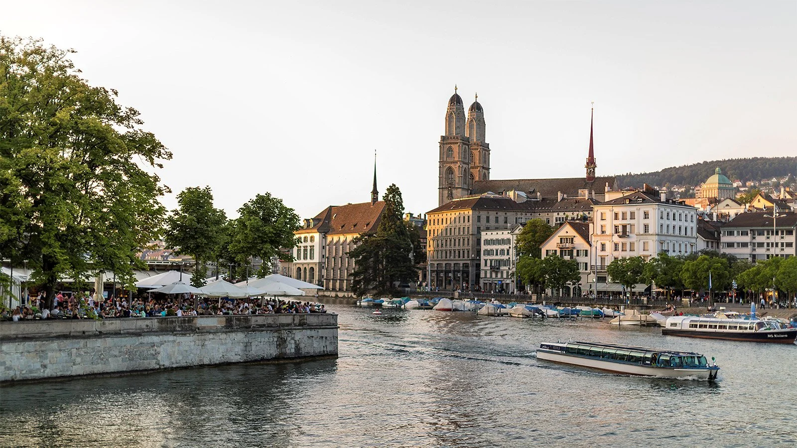 Vue d'une ville avec une rivière, des bateaux, des bâtiments anciens et une grande église gothique en arrière-plan, au coucher du soleil.