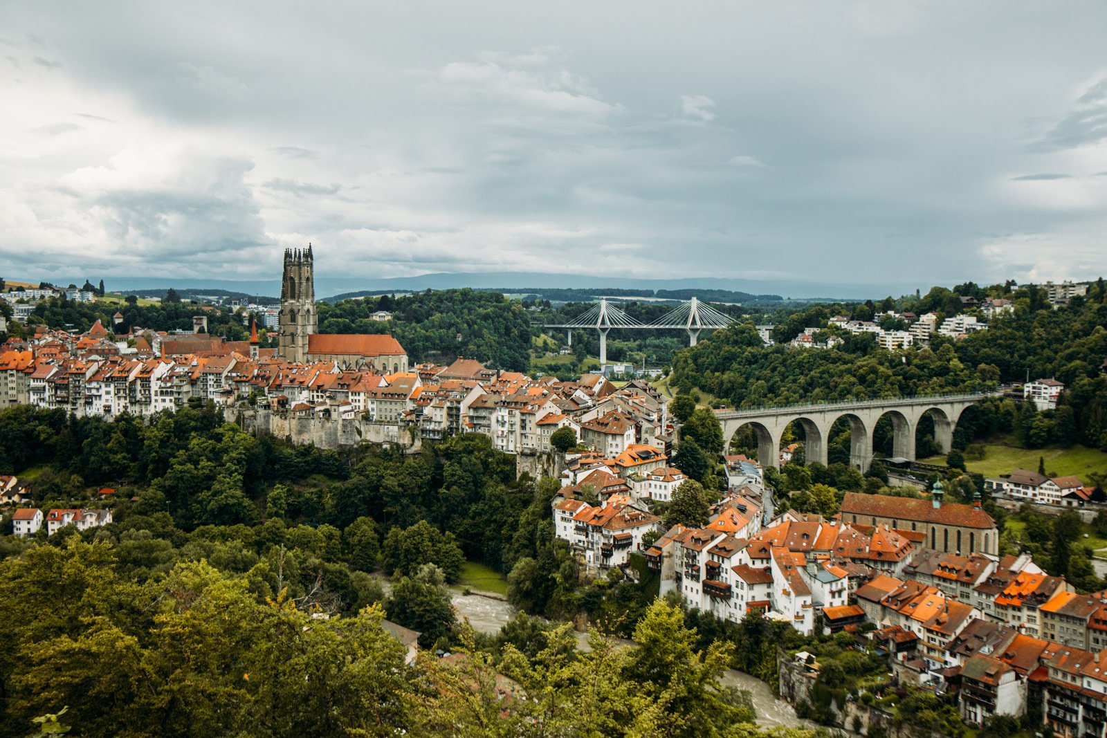 Vue panoramique d'une ville historique avec une église ancienne, des maisons aux toits en tuiles rouges, un pont en arc, et un paysage vallonné avec des ponts modernes, sous un ciel nuageux.