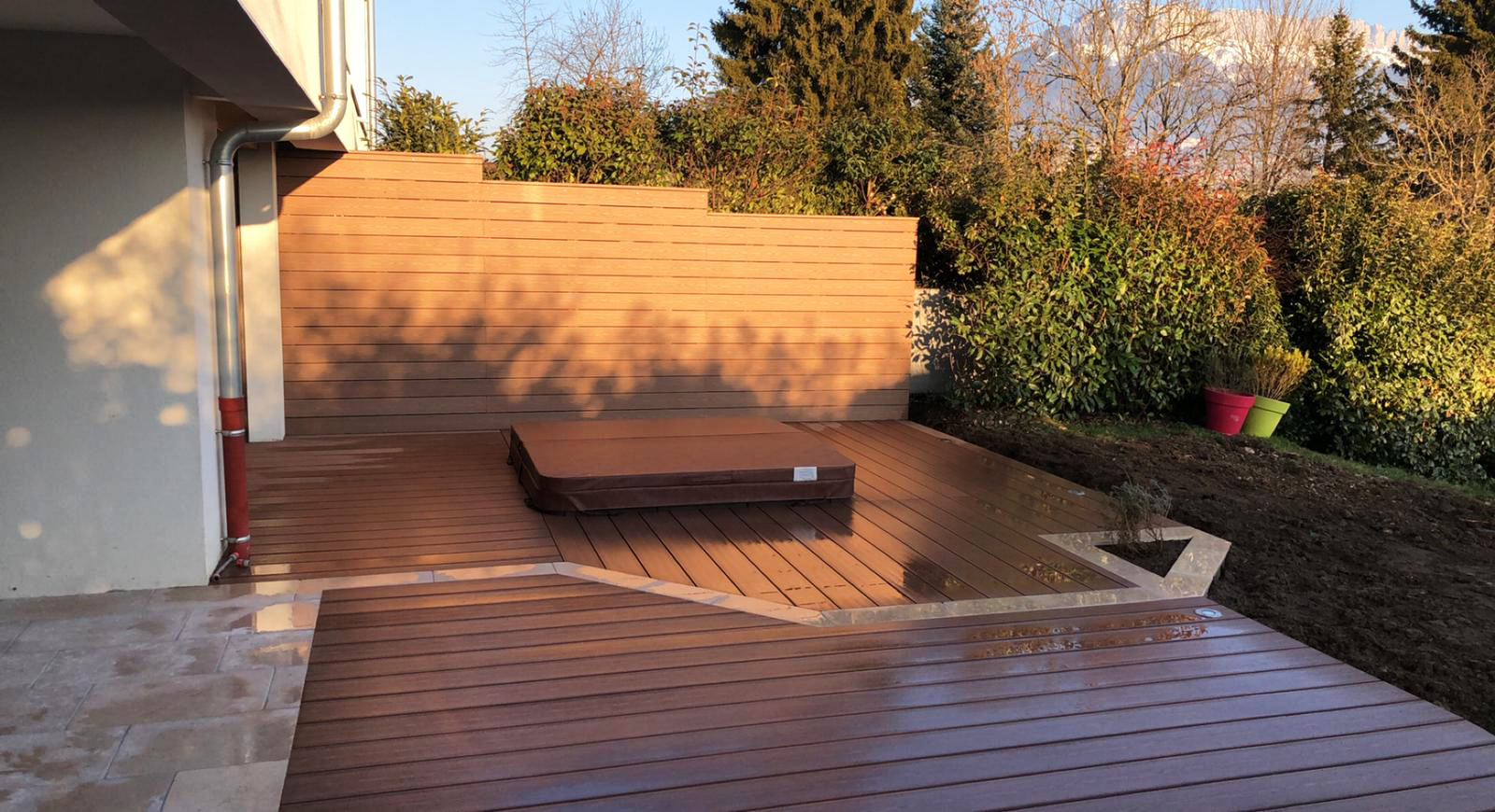 Une terrasse en bois avec un couvercle de jacuzzi, bordée par un panneau d’intimité en bois, avec des buissons et arbres luxuriants en arrière-plan, et deux pots de fleurs vides sur le côté droit.