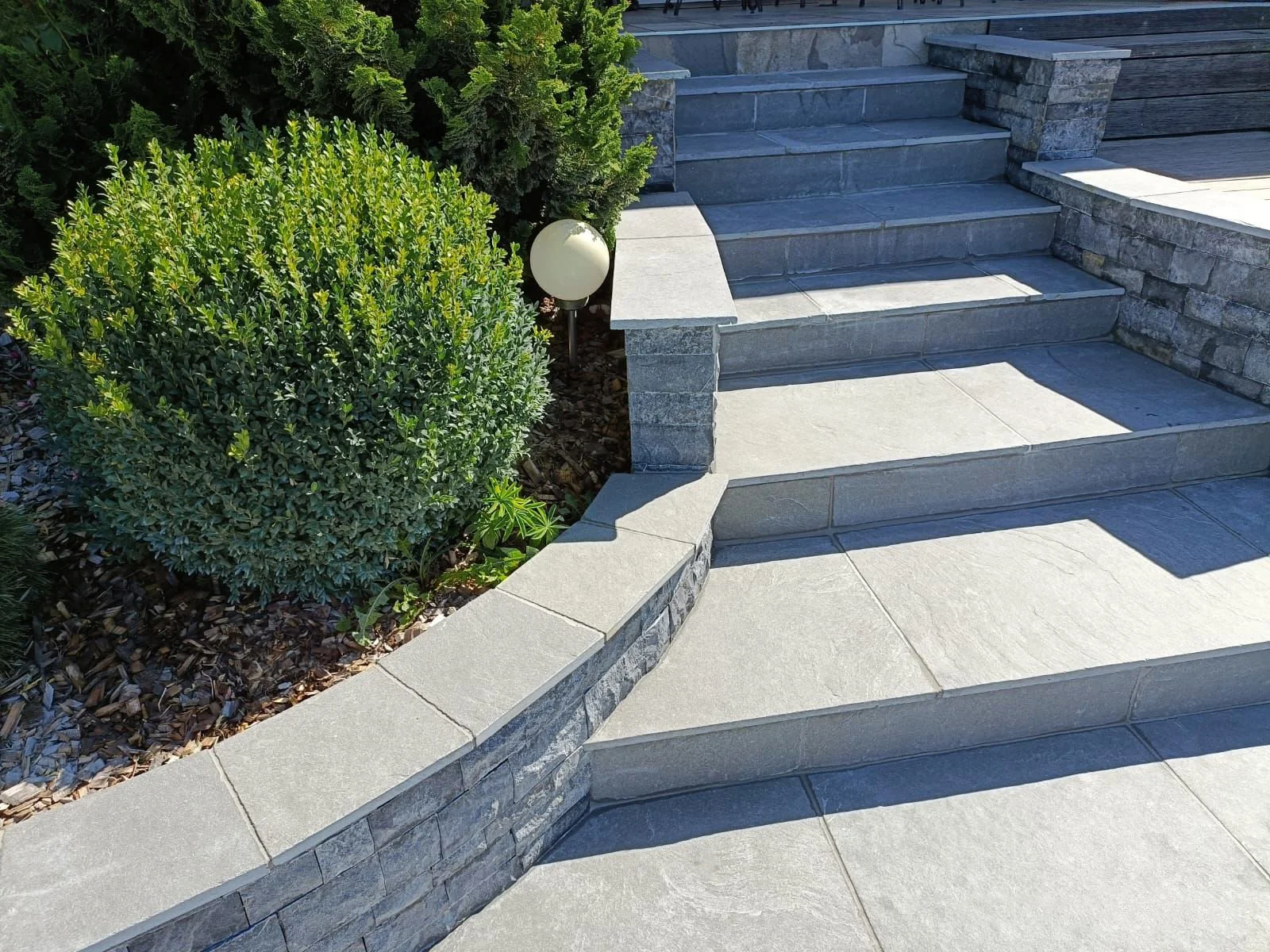 Allée et escalier en pierre, muret en pierre courbé, buissons verts, paillis et lampe extérieure ronde.