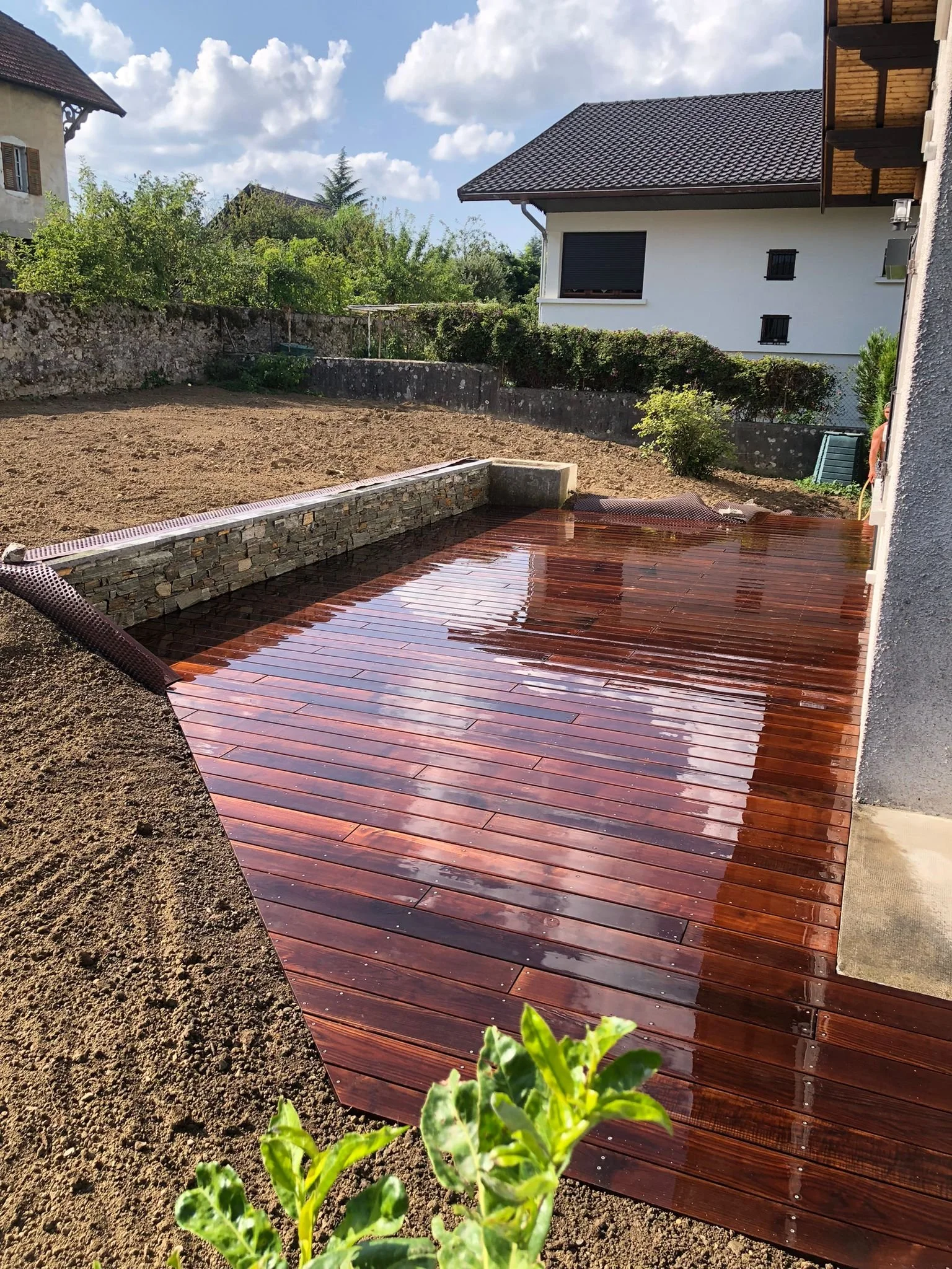 Un jardin arrière avec une terrasse en bois nouvellement installée, entourée de terre et d’un mur de soutènement en pierre.