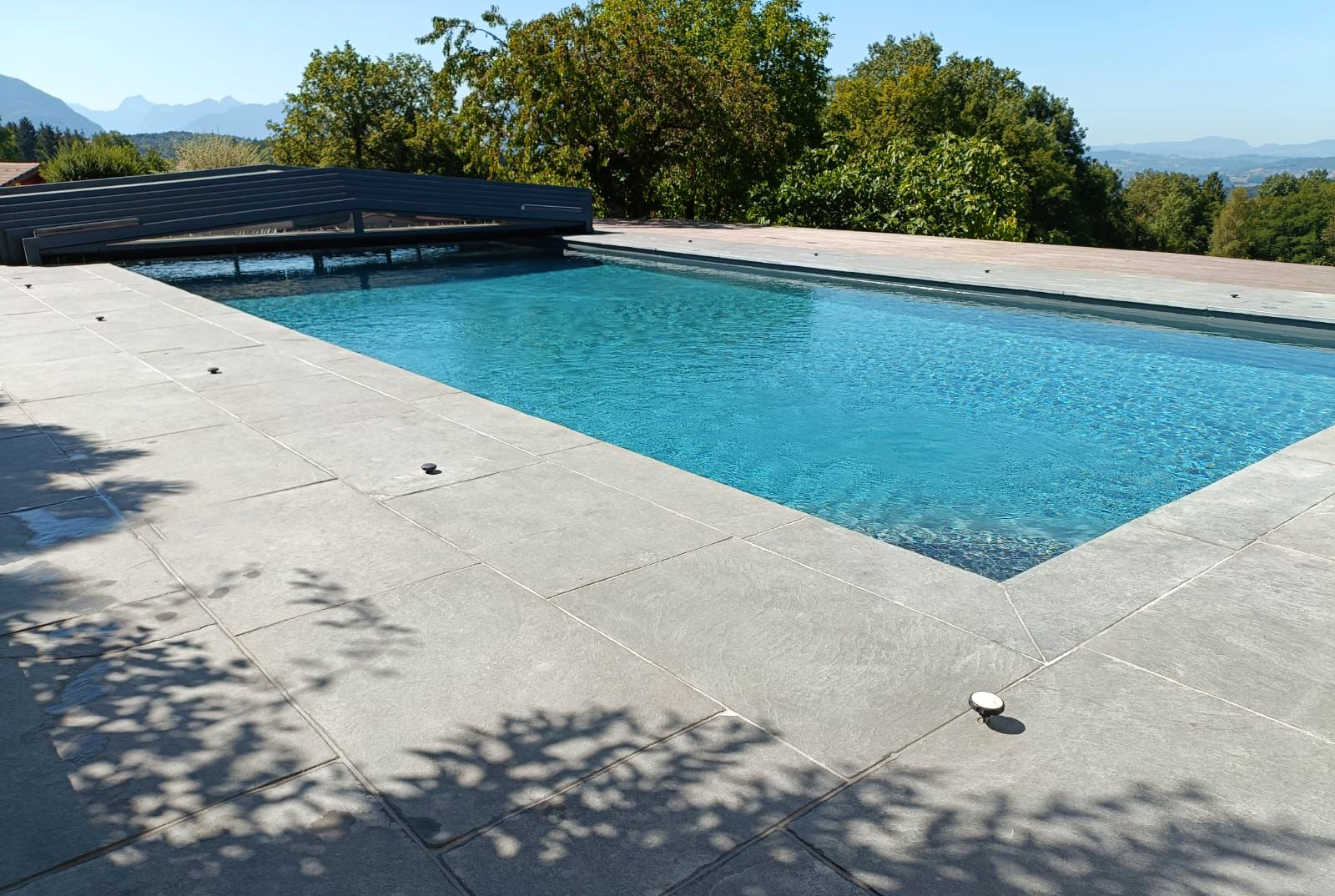 Une piscine rectangulaire avec une terrasse en pierre gris clair, entourée d’arbres verts et de montagnes lointaines sous un ciel bleu clair.