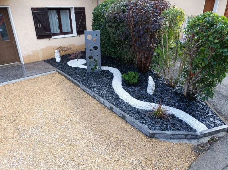 Un massif paysagé avec un chemin sinueux en gravier blanc bordé de pierres noires, entouré de divers arbustes et arbres, avec une sculpture en métal aux découpes circulaires près des plantations.