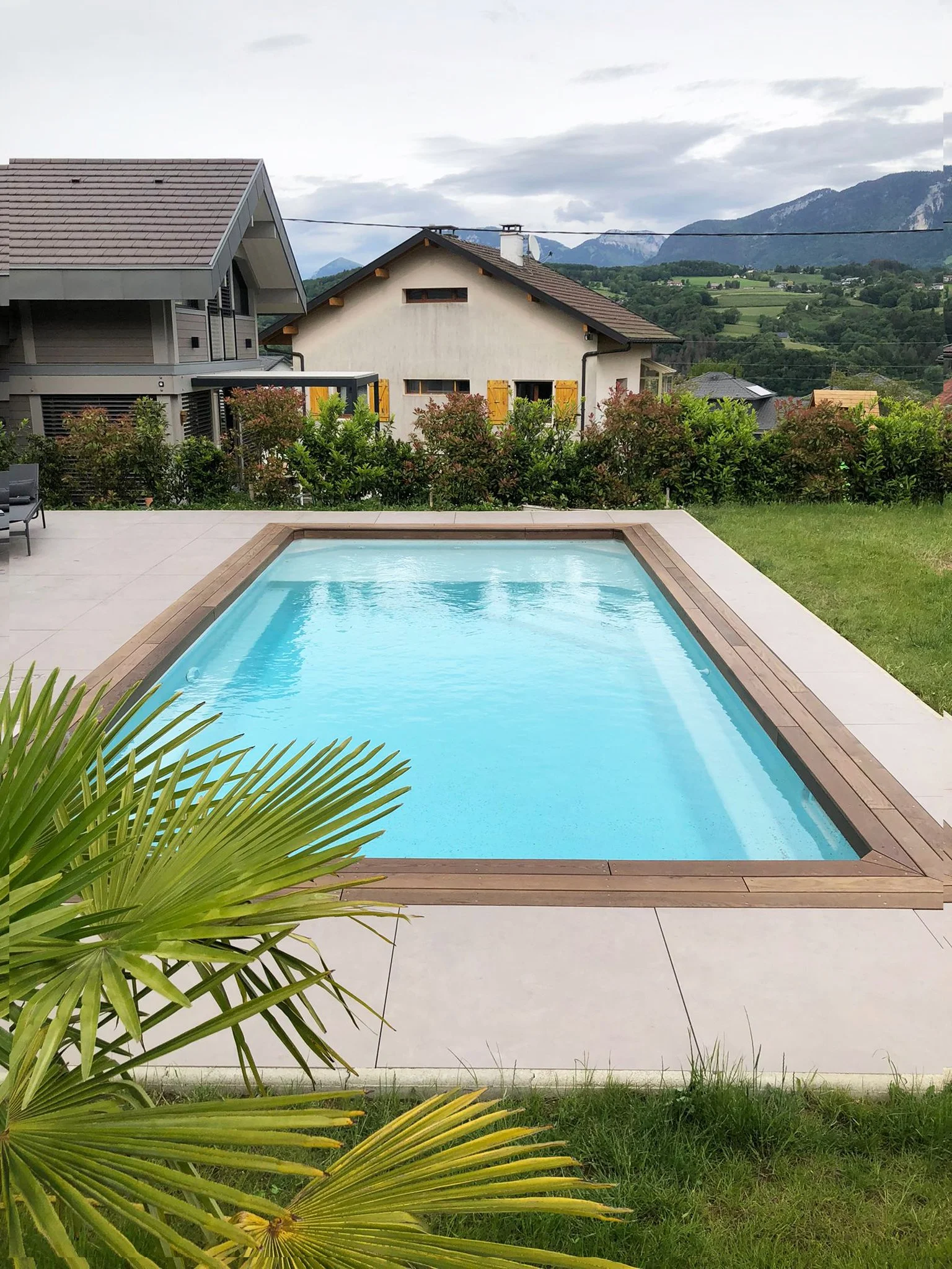 Piscine rectangulaire avec bordure en bois, entourée d’une terrasse carrelée et de verdure, dans un jardin arrière avec des maisons voisines et des montagnes en arrière-plan.