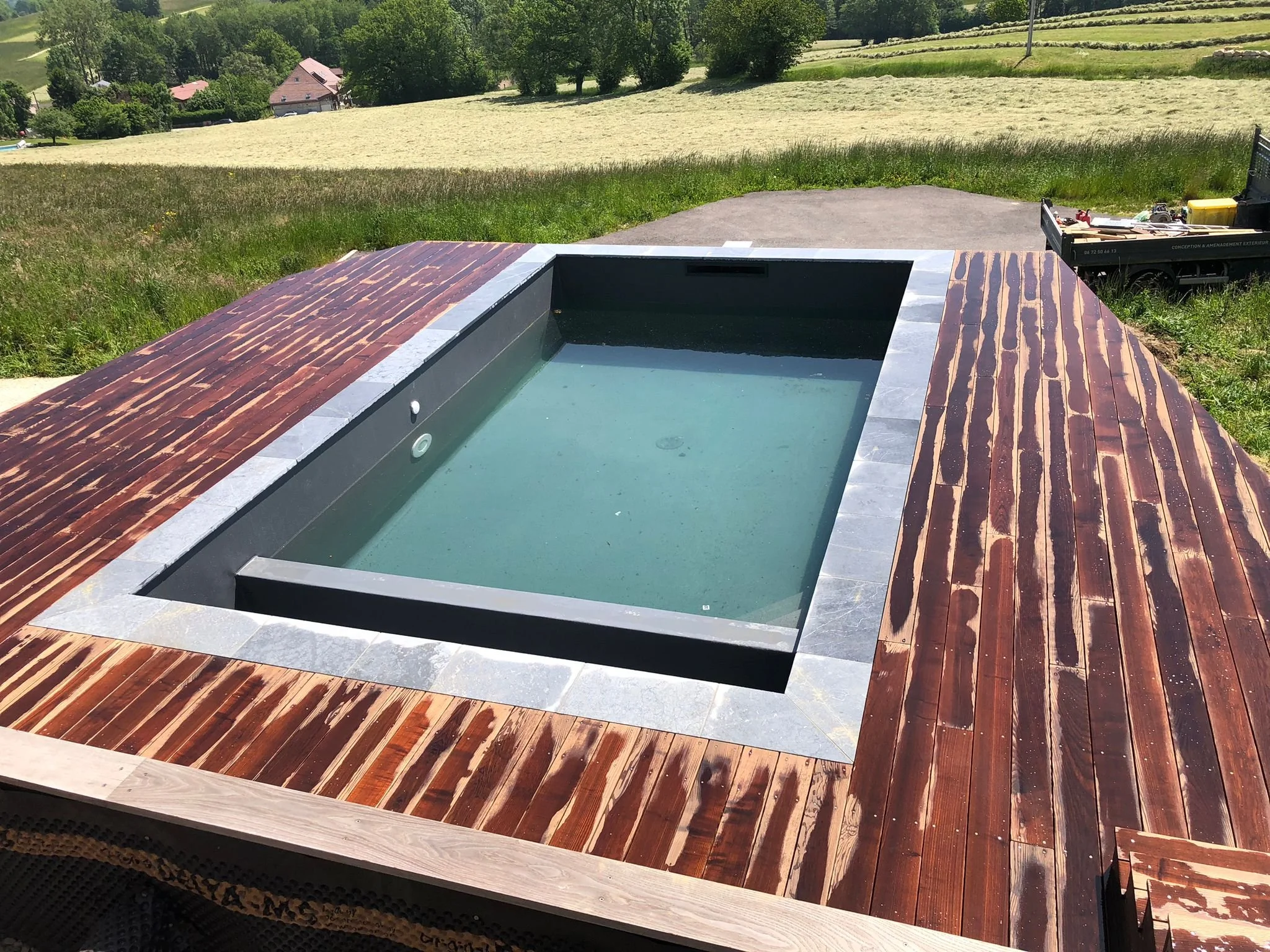 Piscine rectangulaire avec bordure en pierre, entourée d’une terrasse en bois et de verdure.