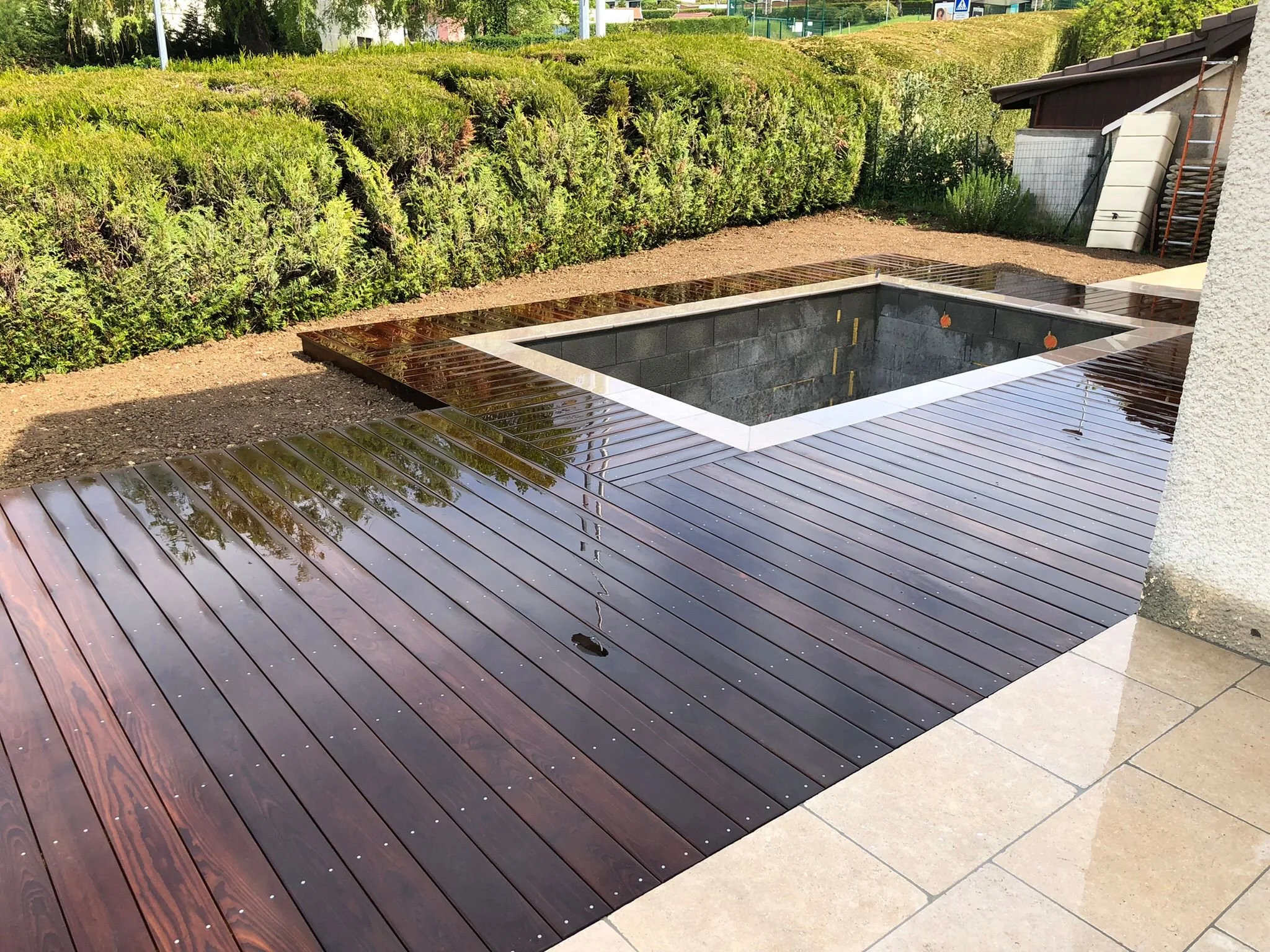 Une piscine entourée d’une terrasse en bois et d’une bordure extérieure blanche, à l’extérieur, près d’une maison avec un sol carrelé beige et des arbustes verts en arrière-plan.
