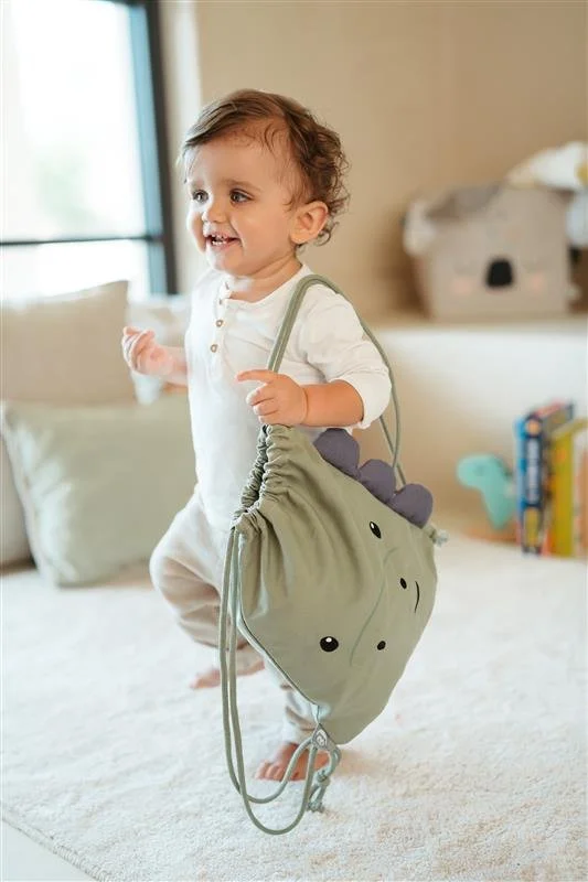 A happy toddler carrying a green dinosaur-themed backpack inside a cozy room with a window, pillow, and children's toys in the background.