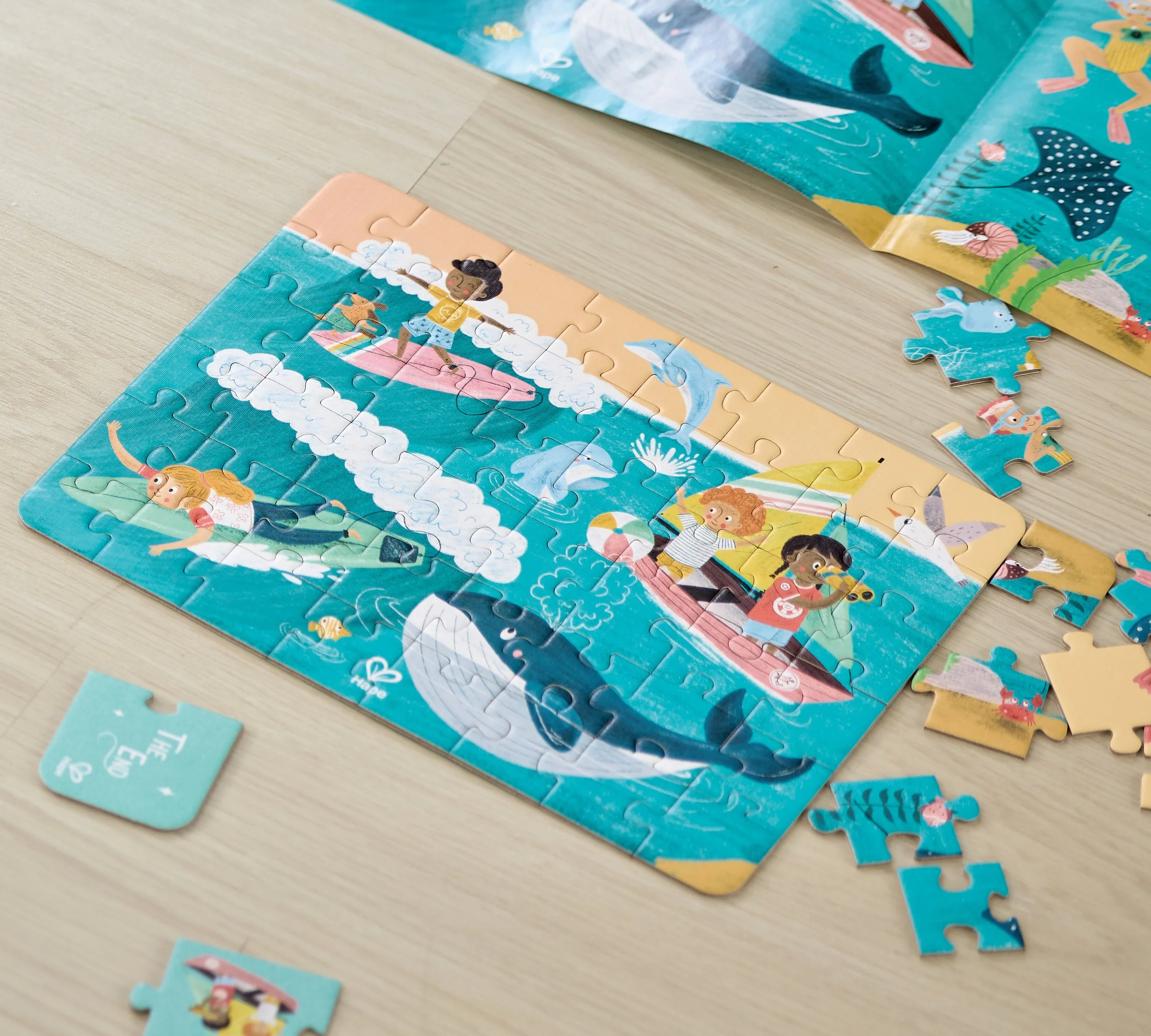 A partially completed Hape puzzle on a light wooden surface, featuring a vibrant underwater scene with children swimming, playing, and dolphins, with loose puzzle pieces scattered around.
