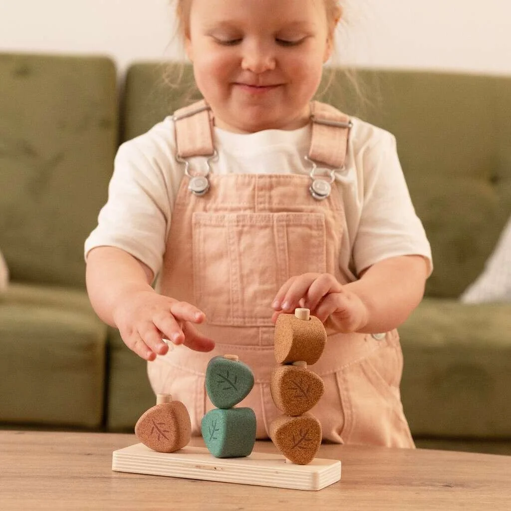 A little girl in a pink jumpsuit stacking cork blocks with leaf carvings on a wooden table. By Korko.