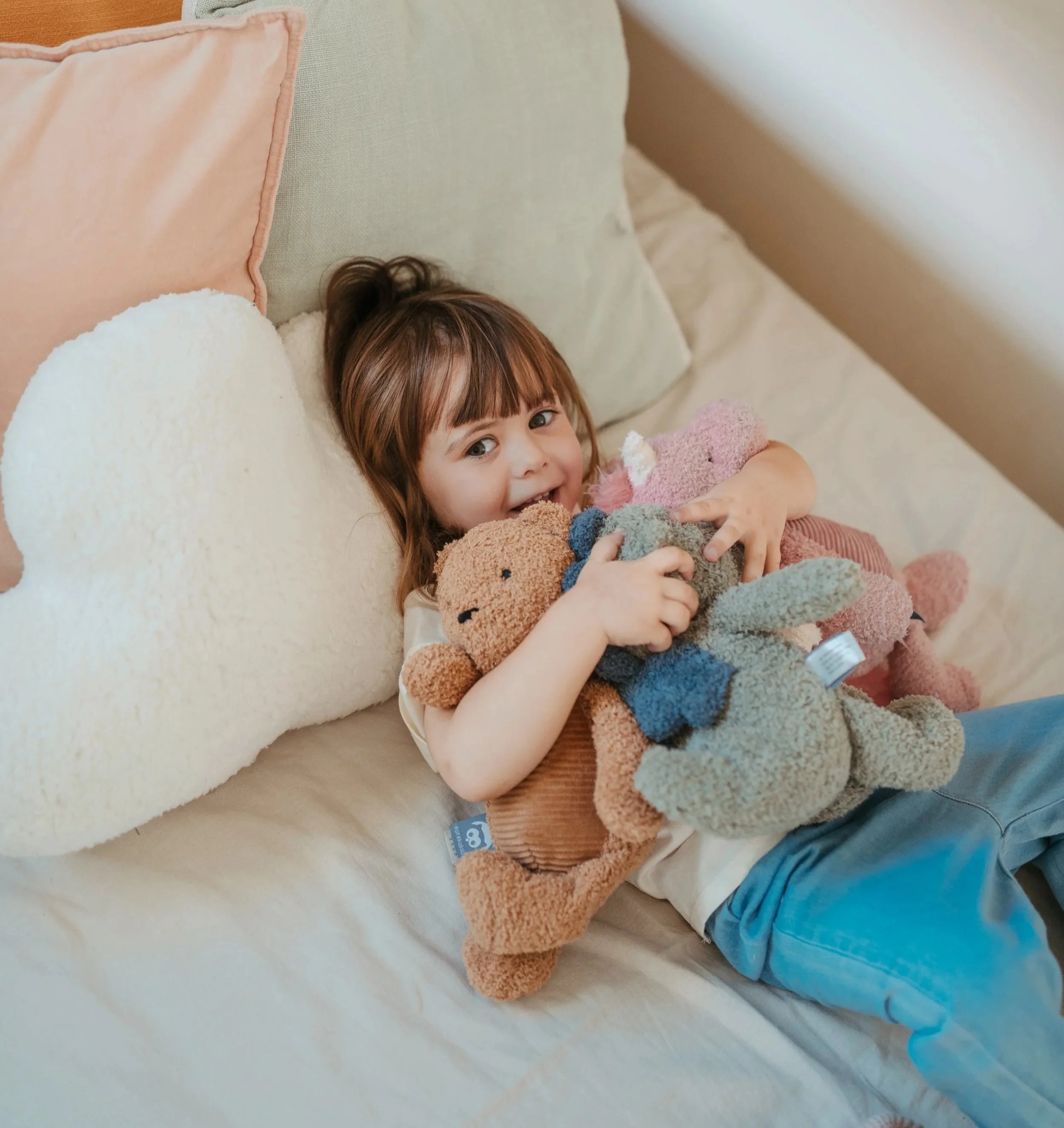 A young girl lying on a bed, hugging several stuffed animals, and smiling at the camera.