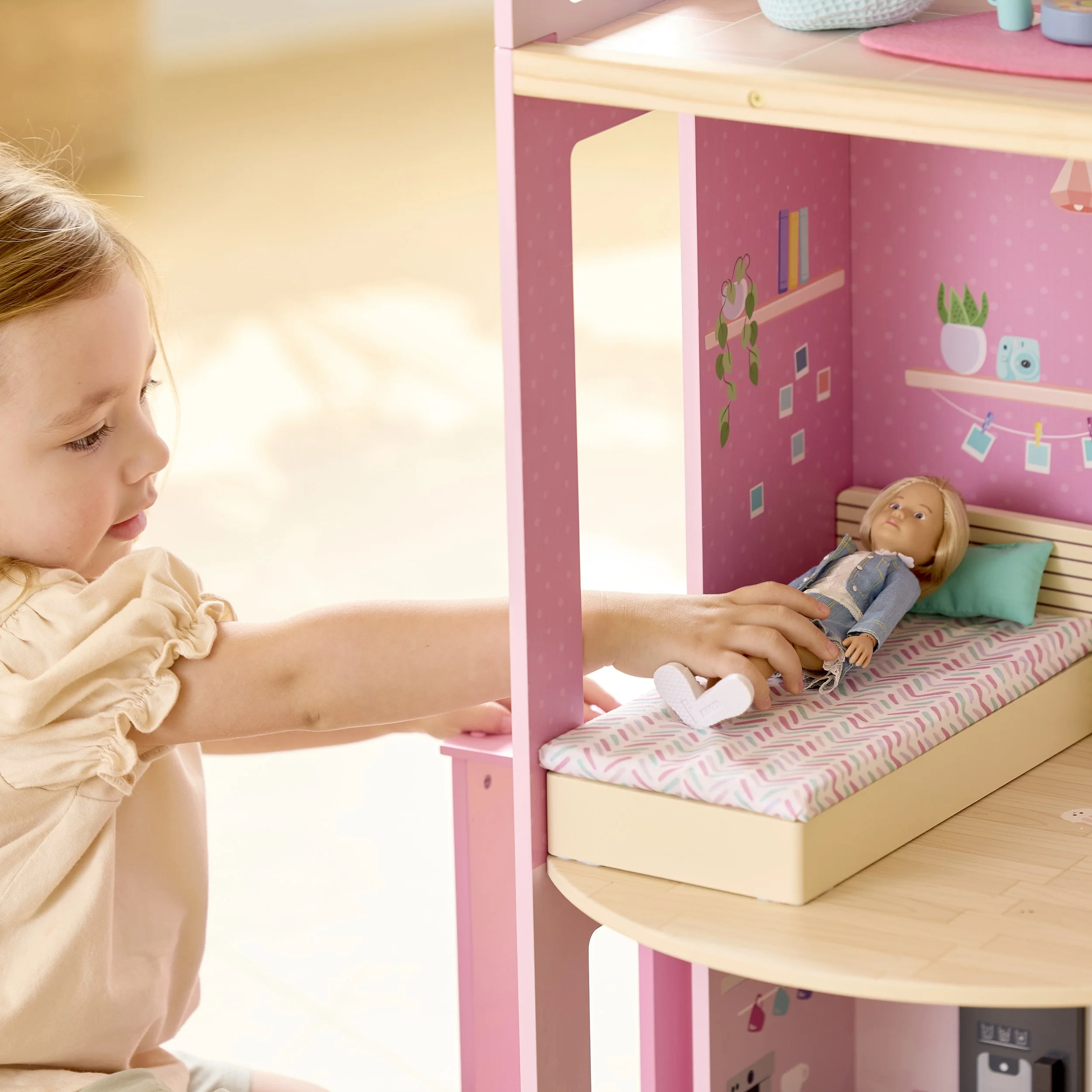 A young girl is playing with a dollhouse, touching a doll lying on a bed inside the dollhouse.