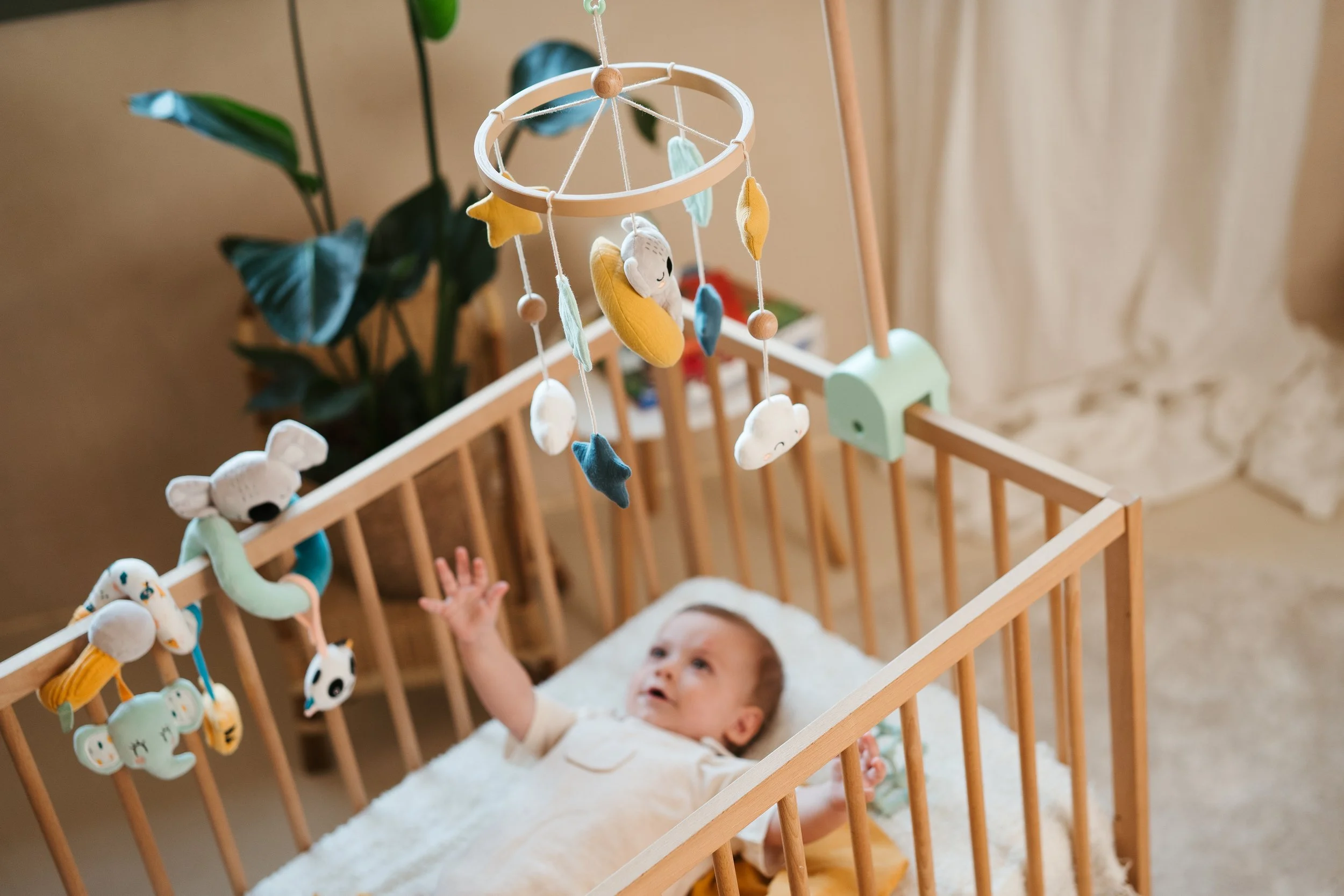 Baby lying in a wooden crib reaching up towards a hanging mobile with plush toys, with a potted plant in the background.