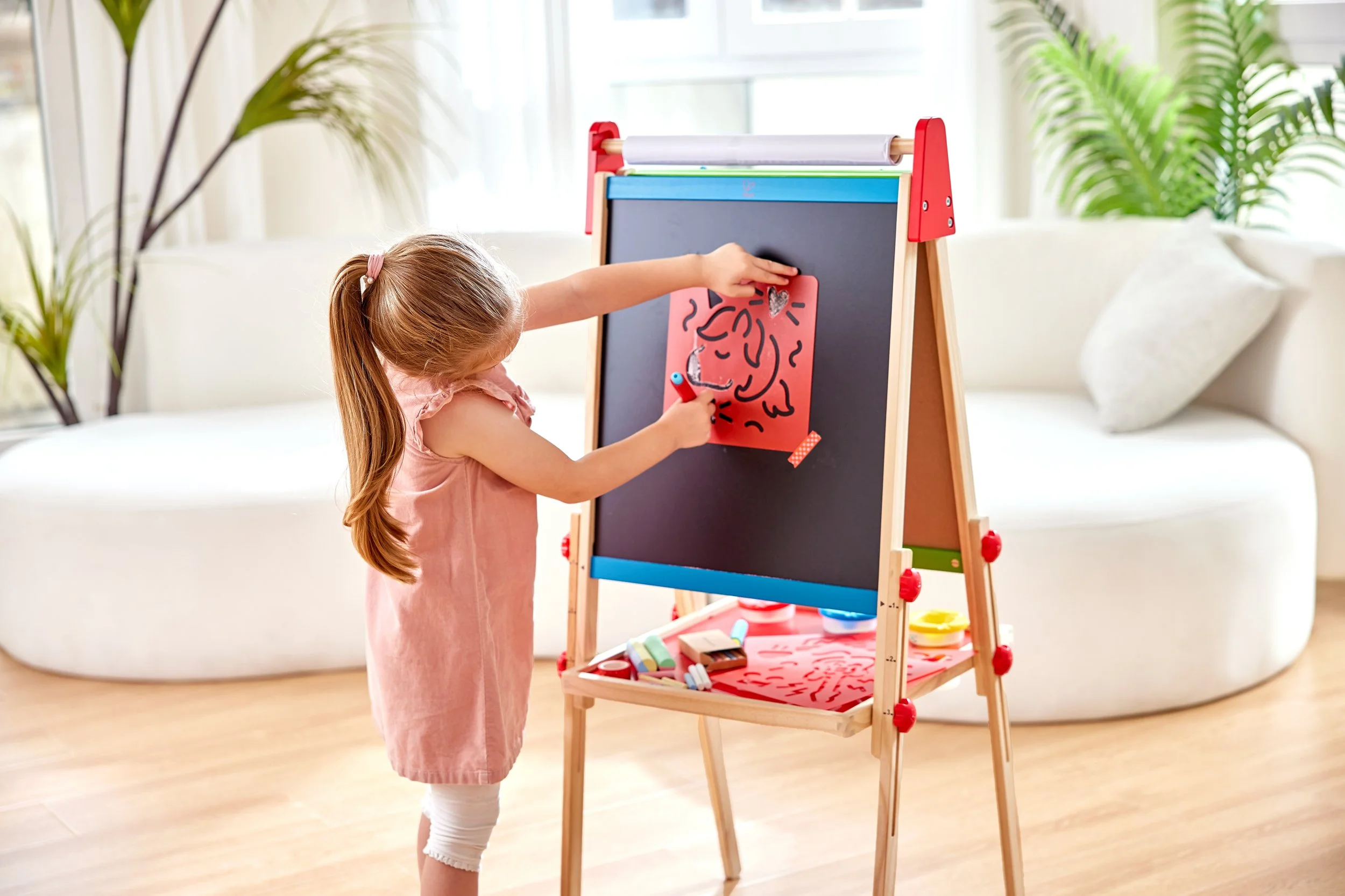 A young girl with long red hair, wearing a pink dress, is drawing on the Hape blackboard easel with a marker and a stencil in a bright, modern living room. The blackboard has a drawing, and there are art supplies on the lower shelf of the easel. Ther