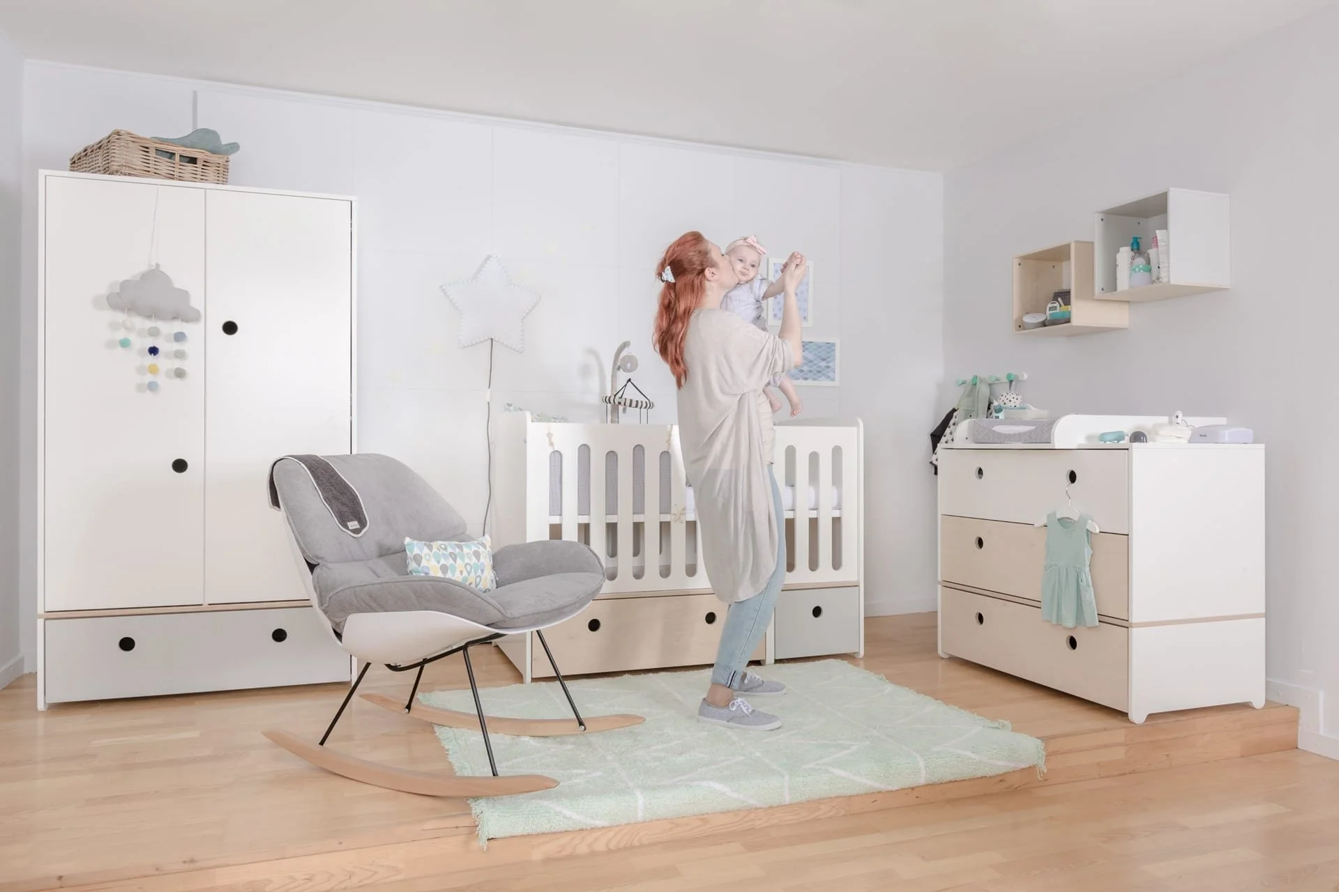 A woman holding a baby in a room with white Colorflex furniture, a gray rocking chair, and light wood flooring.
