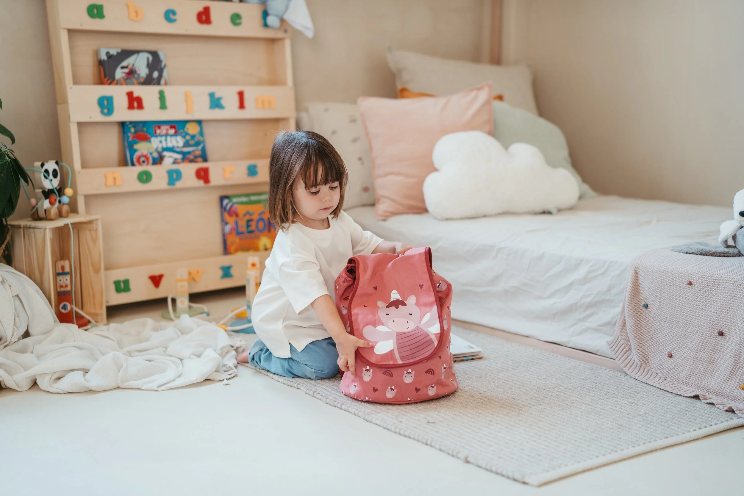 A young girl in a white shirt and blue pants opens a pink backpack with a unicorn illustration in a child's bedroom.