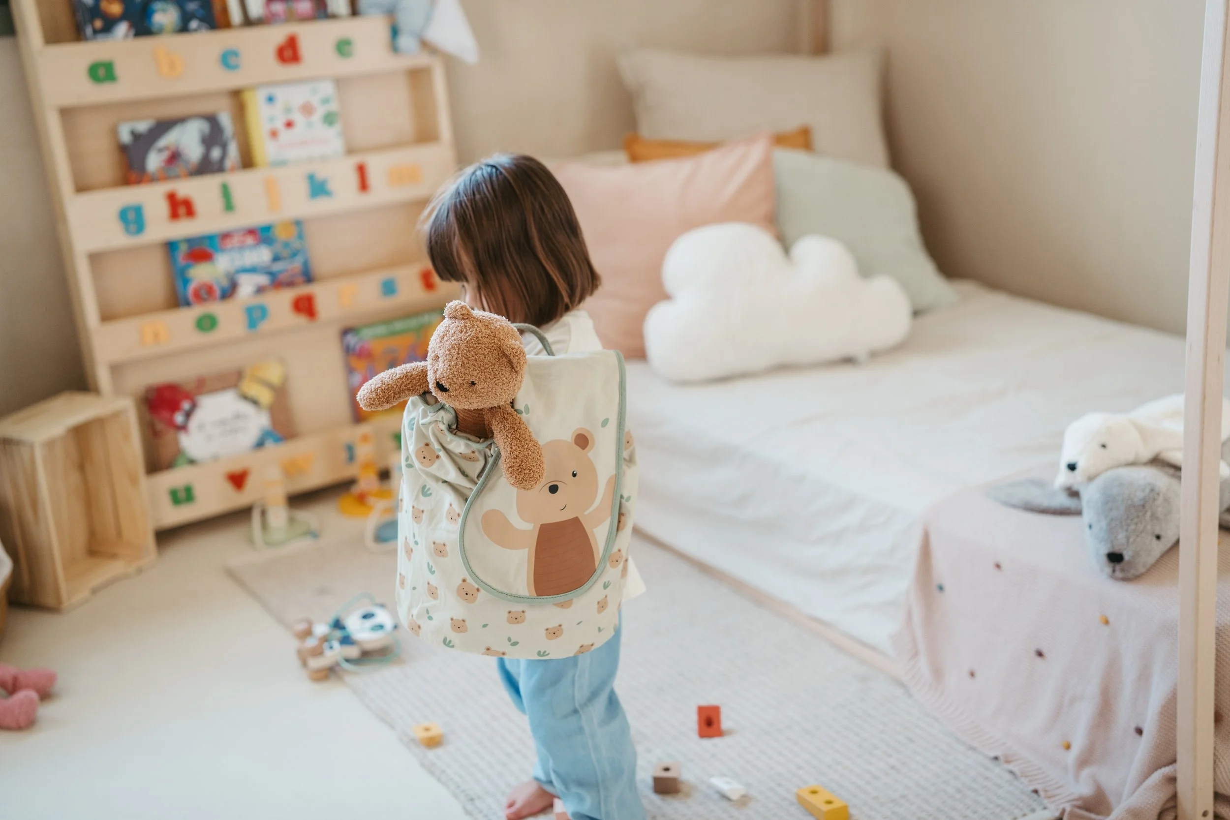 A young child holding a stuffed teddy bear and wearing a backpack with a bear illustration, standing in a bedroom with a bed decorated with cloud and bear plush toys, and a wooden bookshelf filled with colorful books and toys.