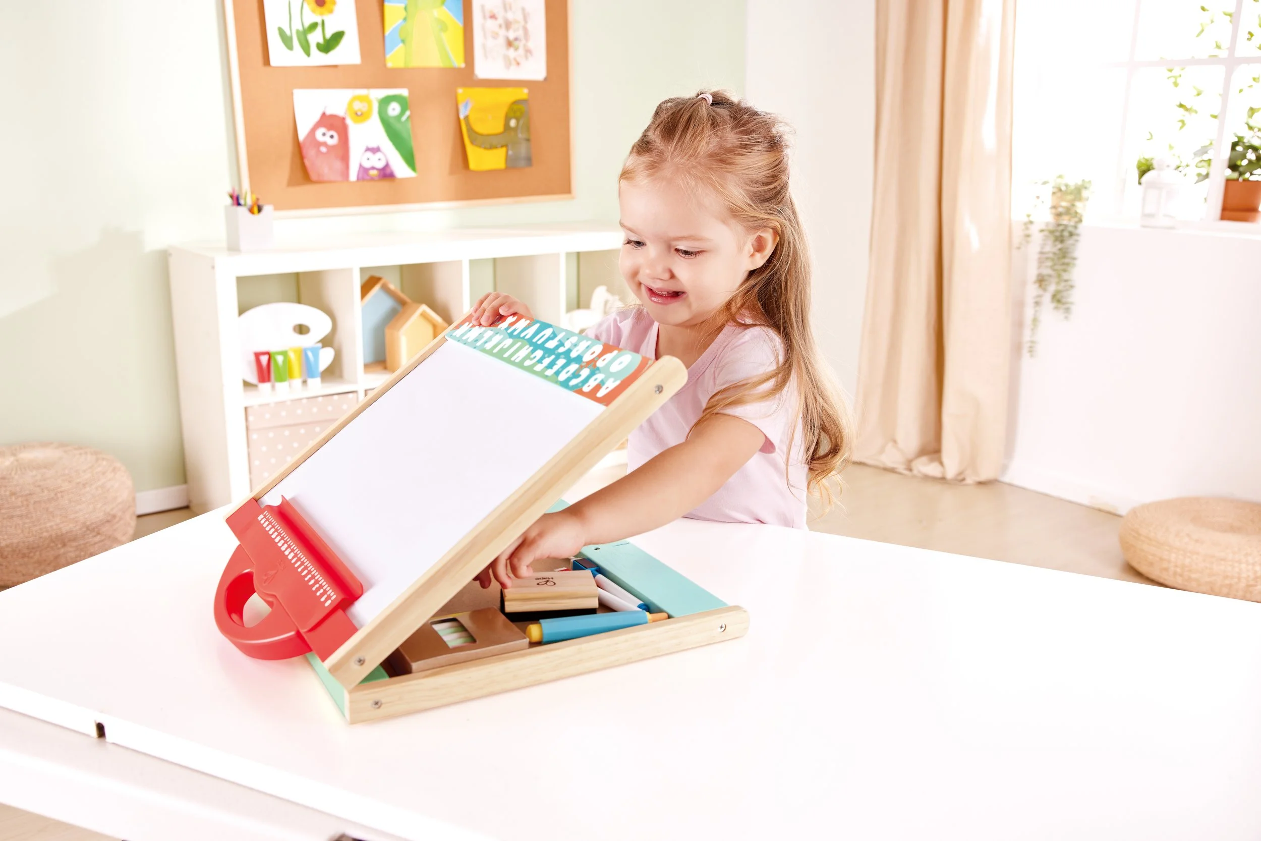 A little girl with long light brown hair gathered in a ponytail is sitting at a white table and opens the magnetic wooden drawing board by Hape with the alphabet and numbers at the top. The room is bright with natural light and decorated with childre