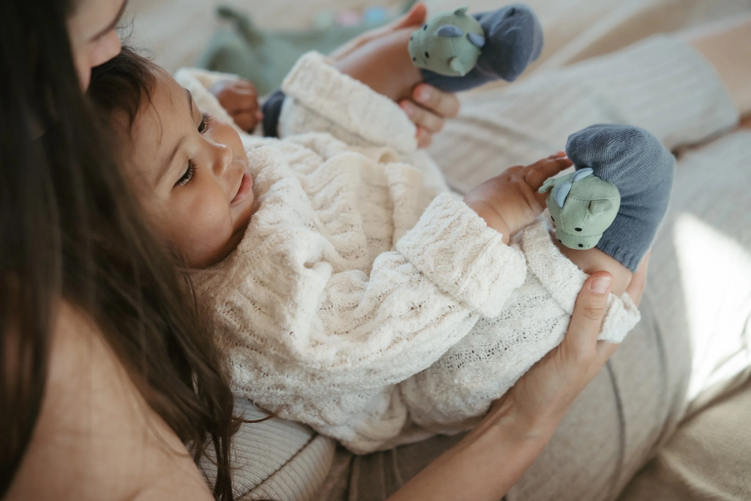 Child lying on a bed, holding plush toys, with an adult nearby.