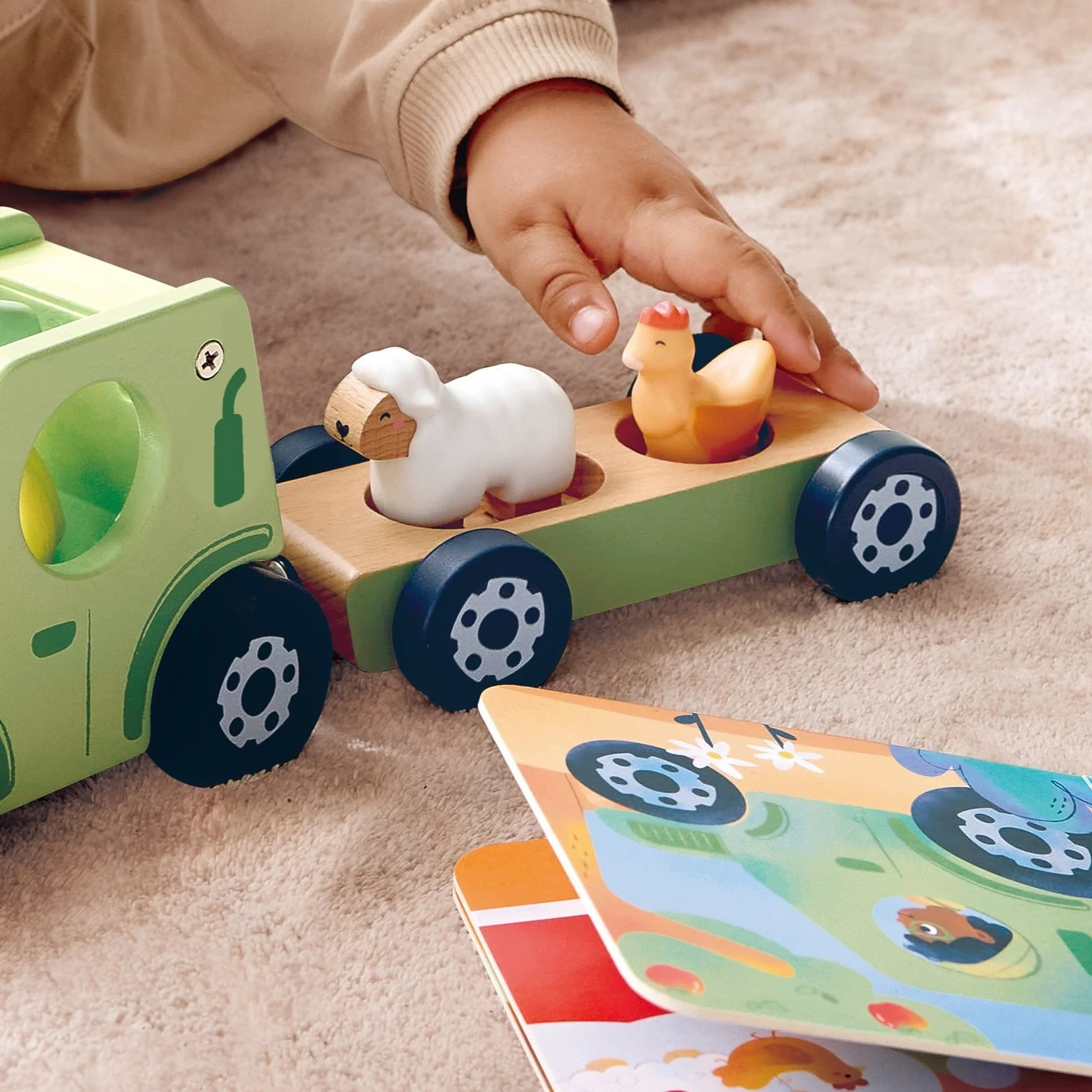 Child's hand placing a sheep and hen figurine on the Hape Land truck, on a beige carpet, with a colorful illustrated book nearby.