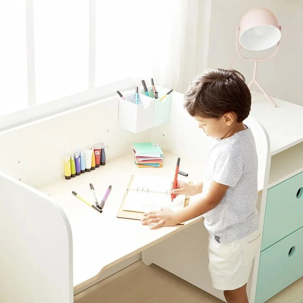 A child in a gray T-shirt and beige shorts standing at a white Colorflex desk, writing in a notebook with various colored markers and pens on the desk, in a bright room with a large window.