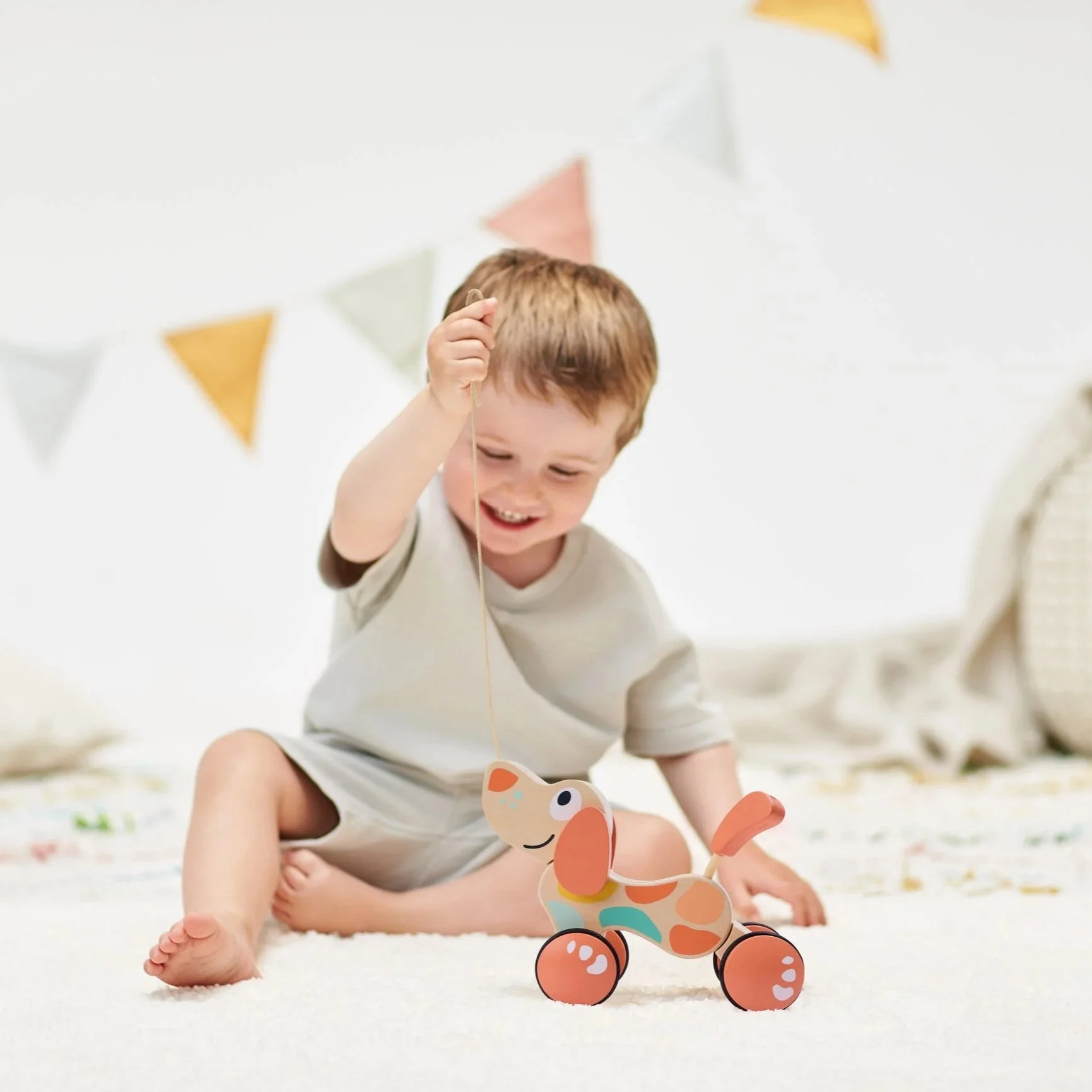 A young boy sitting on a white carpet playing with Hape Pepe pull along special edition for French market in peach and mint colours.