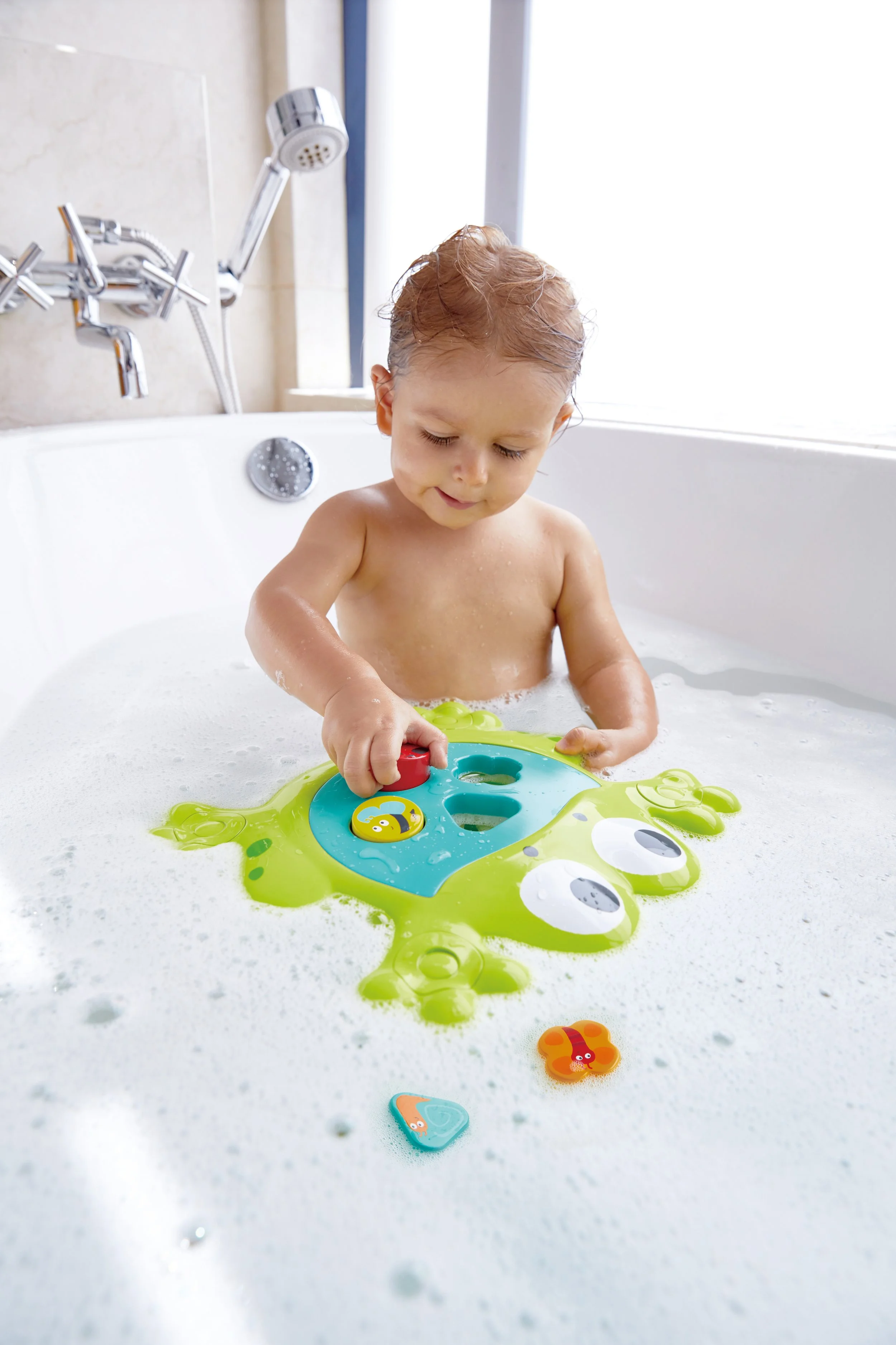 A young child playing in a bathtub with "Feed-me bath frog" by Hape, surrounded by bubbles, with a window in the background.