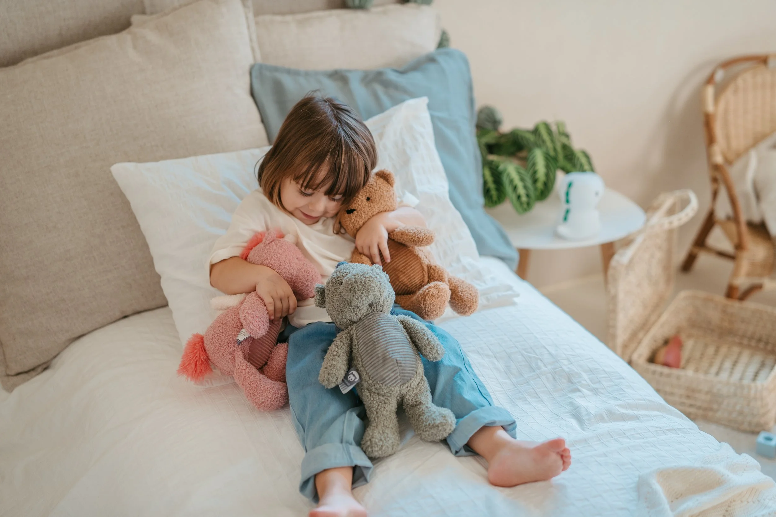 A young girl with short brown hair lying on a bed with white bedding, holding and playing with three teddy bears, surrounded by pillows and cozy decor.