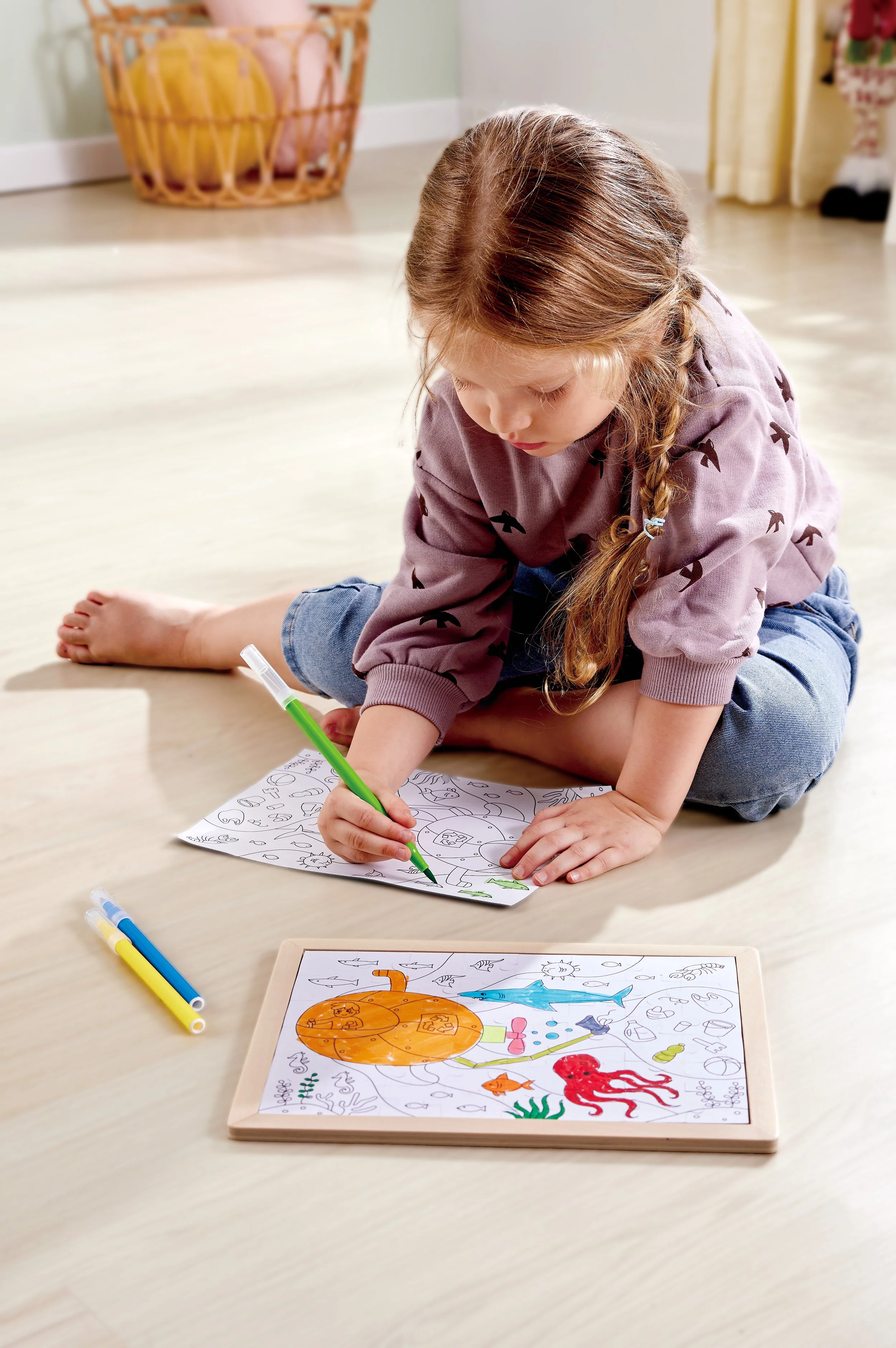 A young girl sitting on the floor decorating a d coloring sheet with colored markers, with a completed coloring page of a dolphin, octopus, and various sea creatures in front of her.