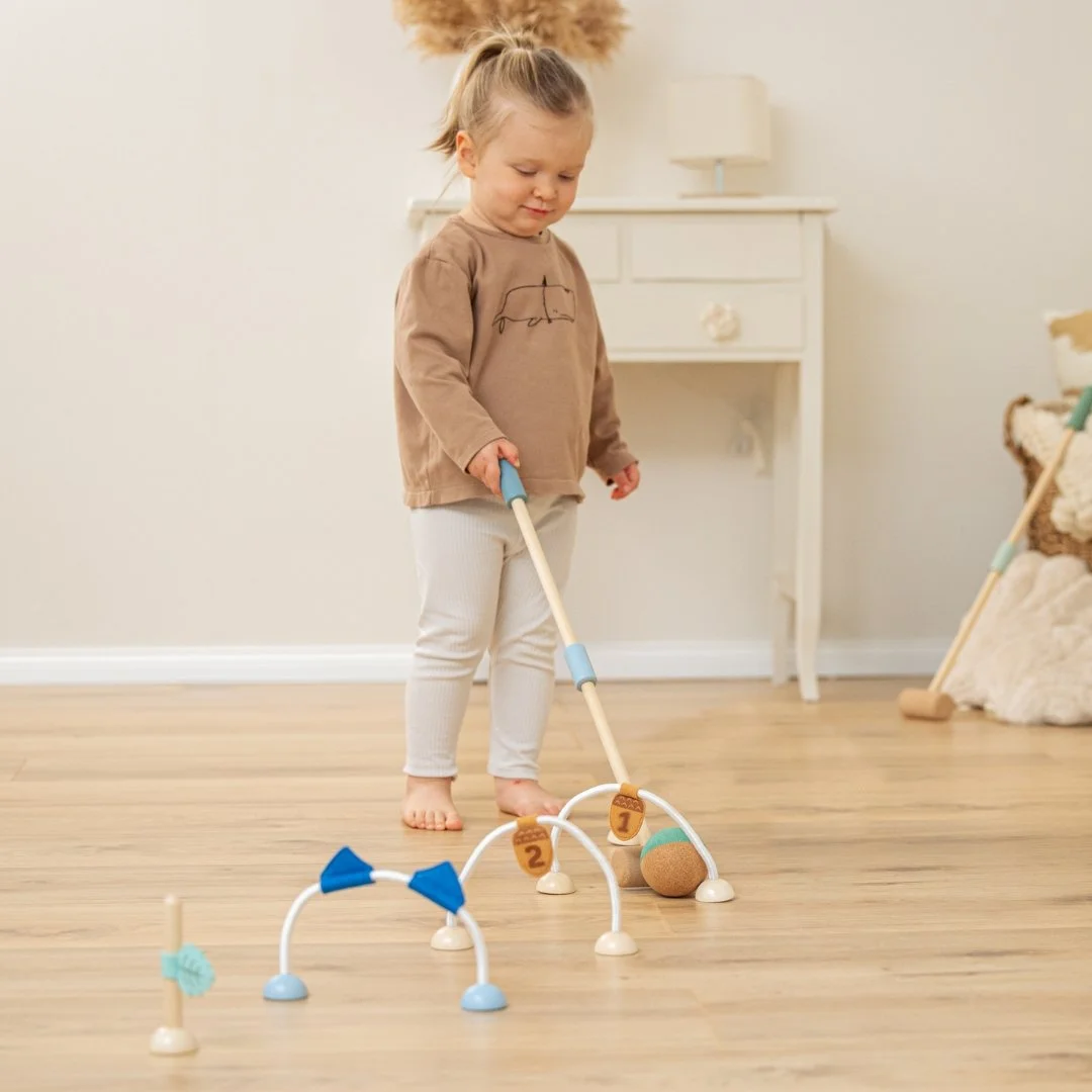 A little girl plays golf indoors on a wooden floor, holds a toy golf club and is about to hit cork golf balls at plastic bows with numbered labels. By Korko.