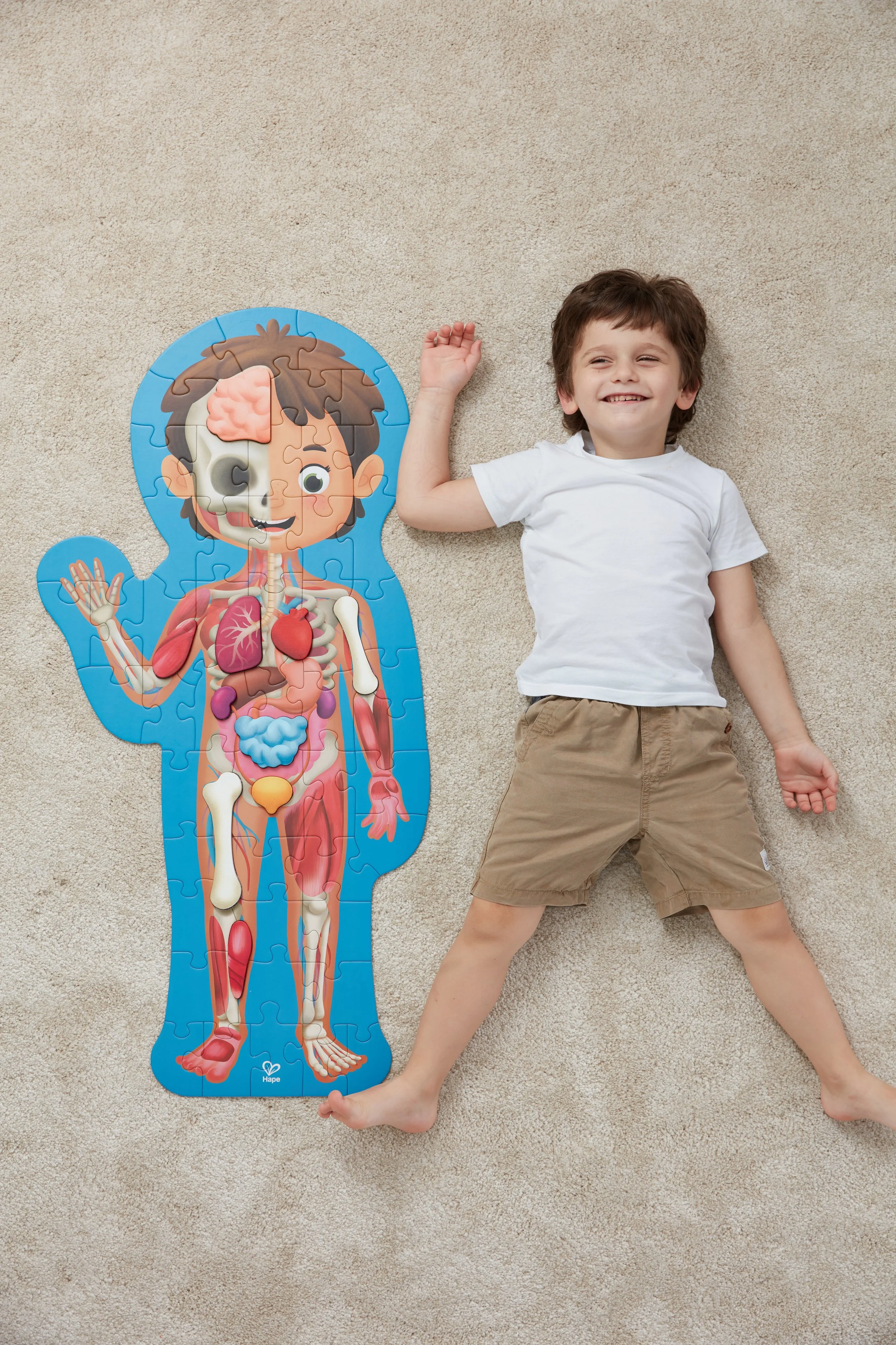 A young boy lying on a beige carpet next to a large Hape jigsaw puzzle of a human body's internal organs, smiling and waving.