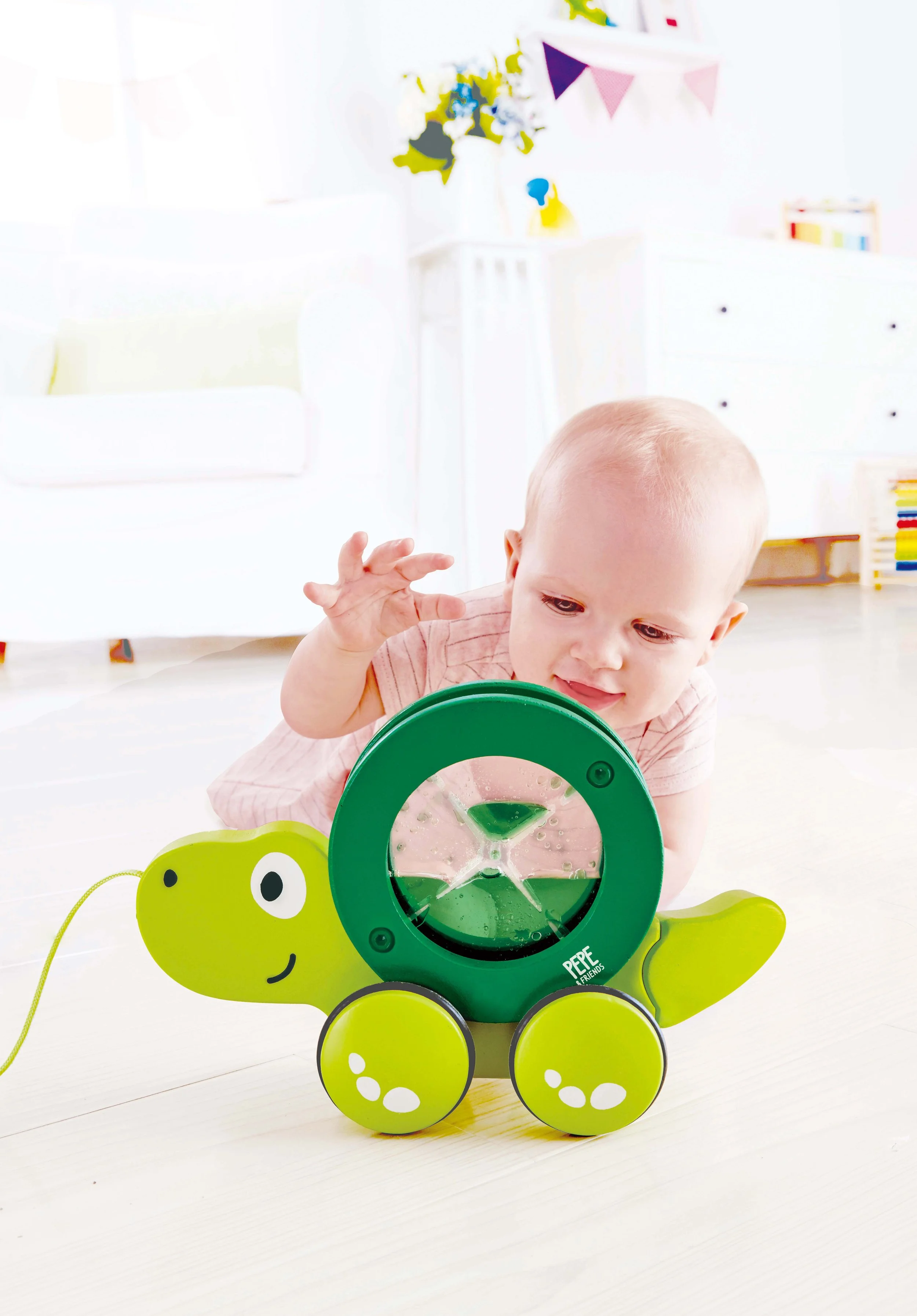 A baby lying on the floor, playing with a green toy shaped like a turtle with wheels, in a bright, white room decorated with bunting and flowers. Developed by Hape.