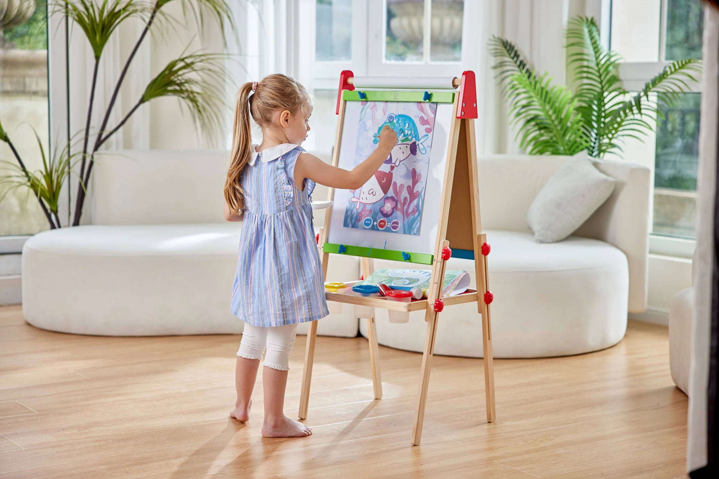 A young girl with long hair tied in a ponytail wearing a blue and white striped dress and white leggings drawing and coloring a poster on an easel in a bright living room filled with natural light and green plants.