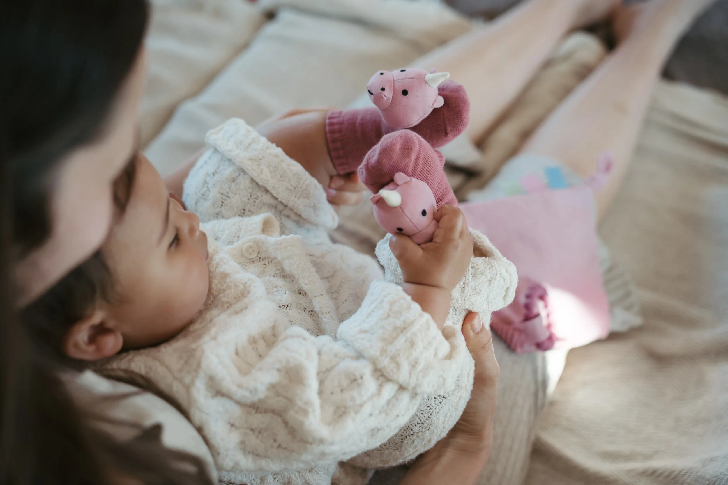 A young girl lying on a bed, holding two pink stuffed animal pigs with white horns, dressed in pink hats. She is gazing at the toys, which are close to her face.