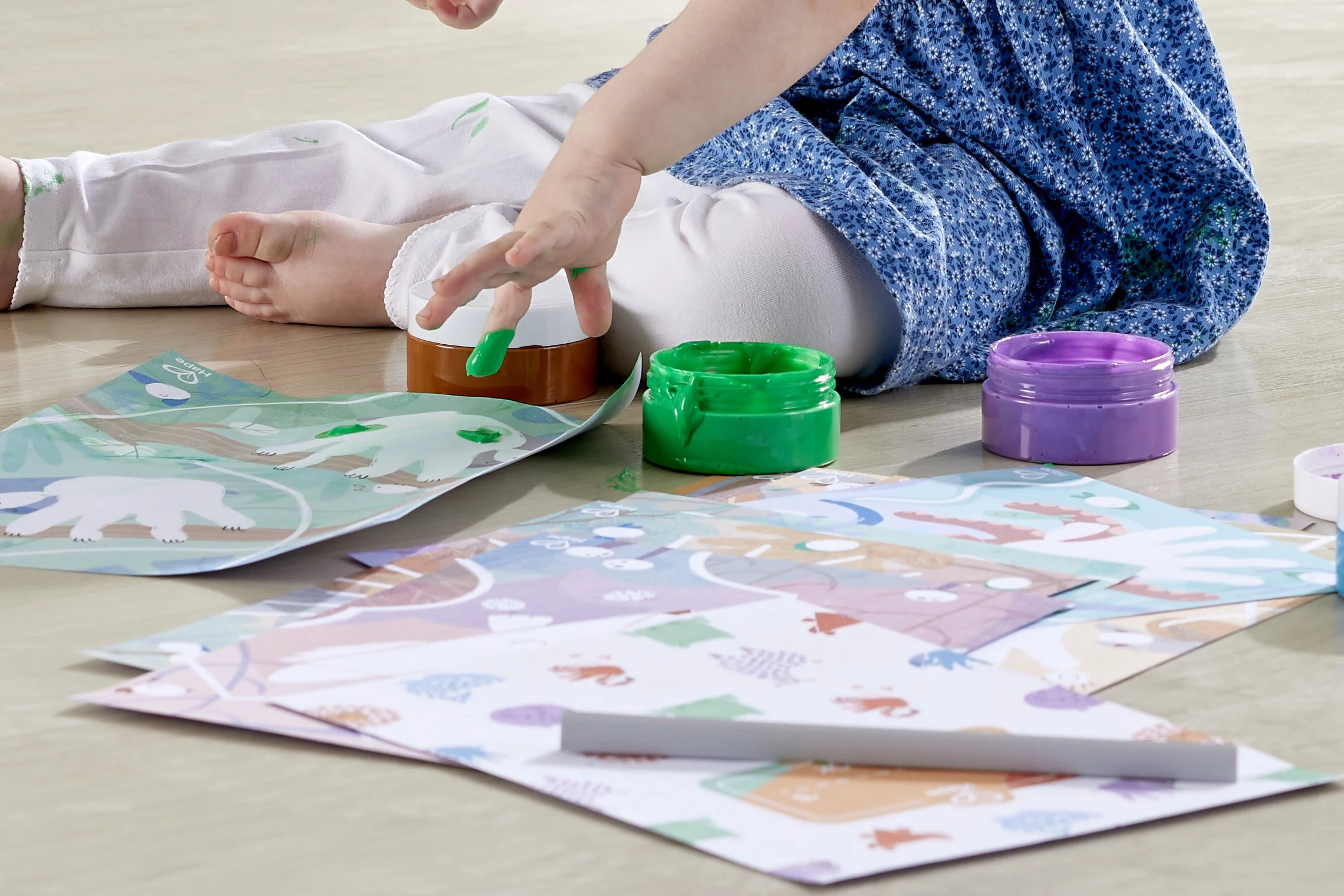 Child painting with fingers on paper with green paint, surrounded by open paint pots in orange, green, and purple, on a wooden floor.