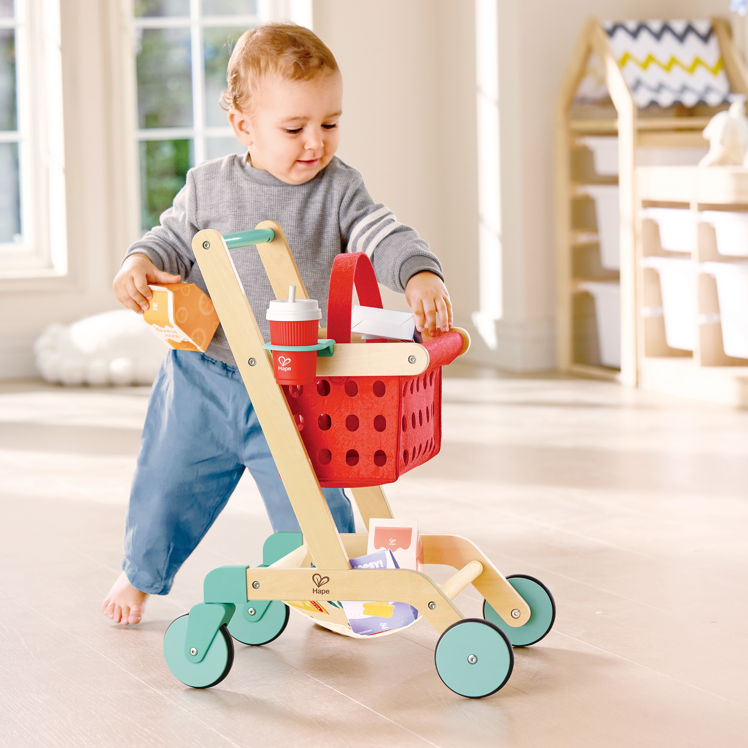 A young child playing with a toy shopping cart filled with toy groceries in a bright room with wooden flooring and large windows.