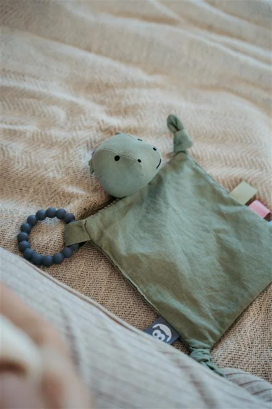 A plush toy with a round head and small face, attached to a soft fabric blankie with a beaded loop. The items are on a textured beige surface, possibly a blanket or bedspread.