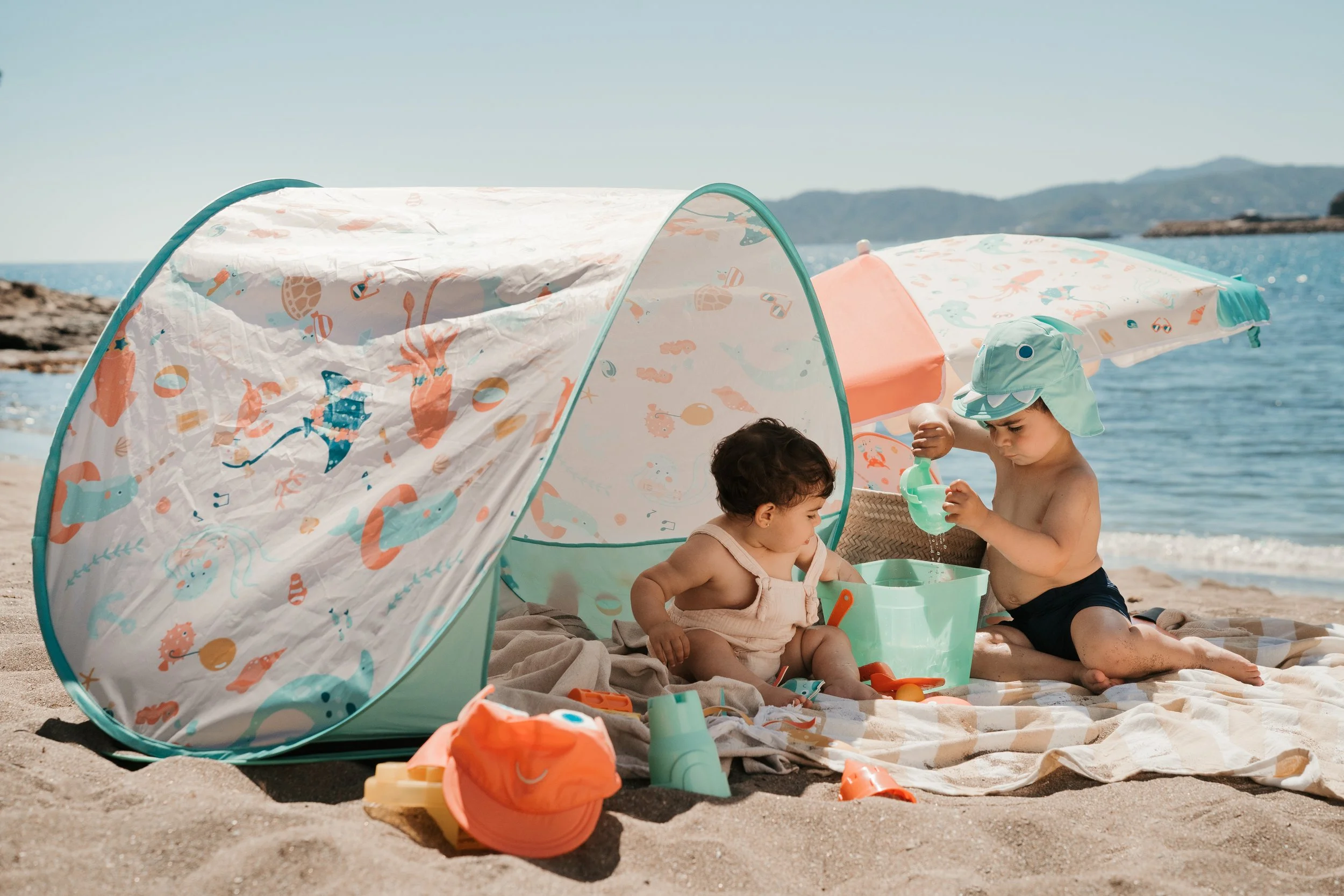 Two young children playing in a sandpit on the beach, under a colorful beach tent from the Eurekakids Beach Collection, with ocean and distant hills in the background.